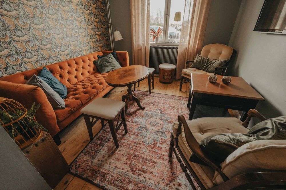 A cozy living room corner with a brown leather Chesterfield sofa, exposed brick wall, and vintage-style decor.