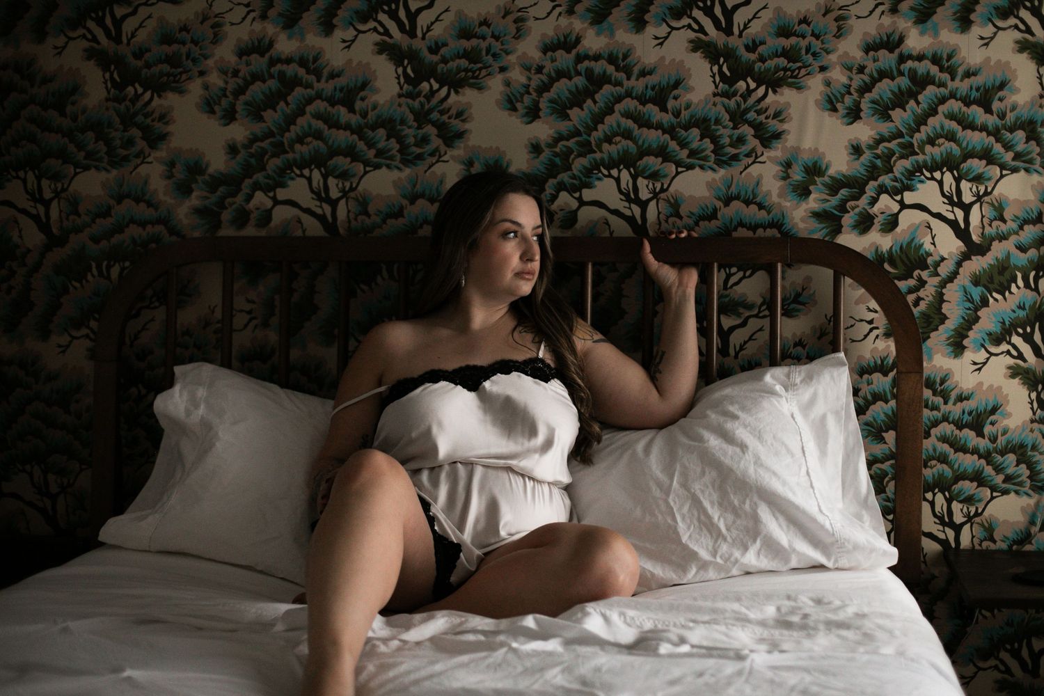 A person in a white strapless dress poses elegantly on a bed with decorative wallpaper in the background.