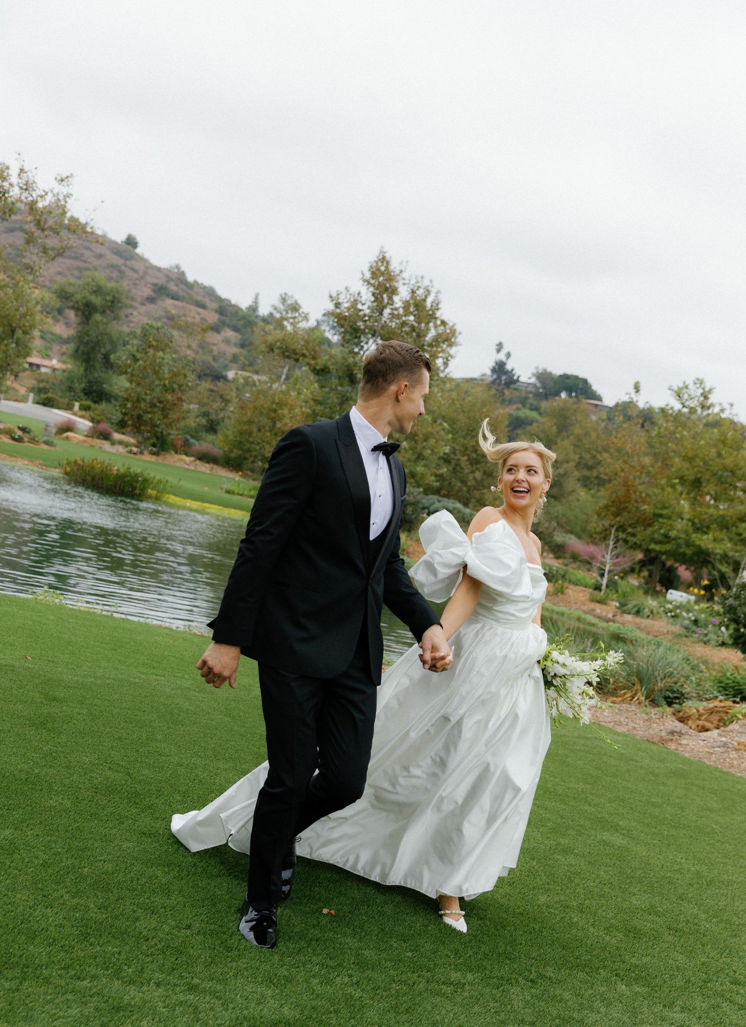 A joyful couple runs together on a lush green lawn by a lake during their wedding celebration at an outdoor venue.