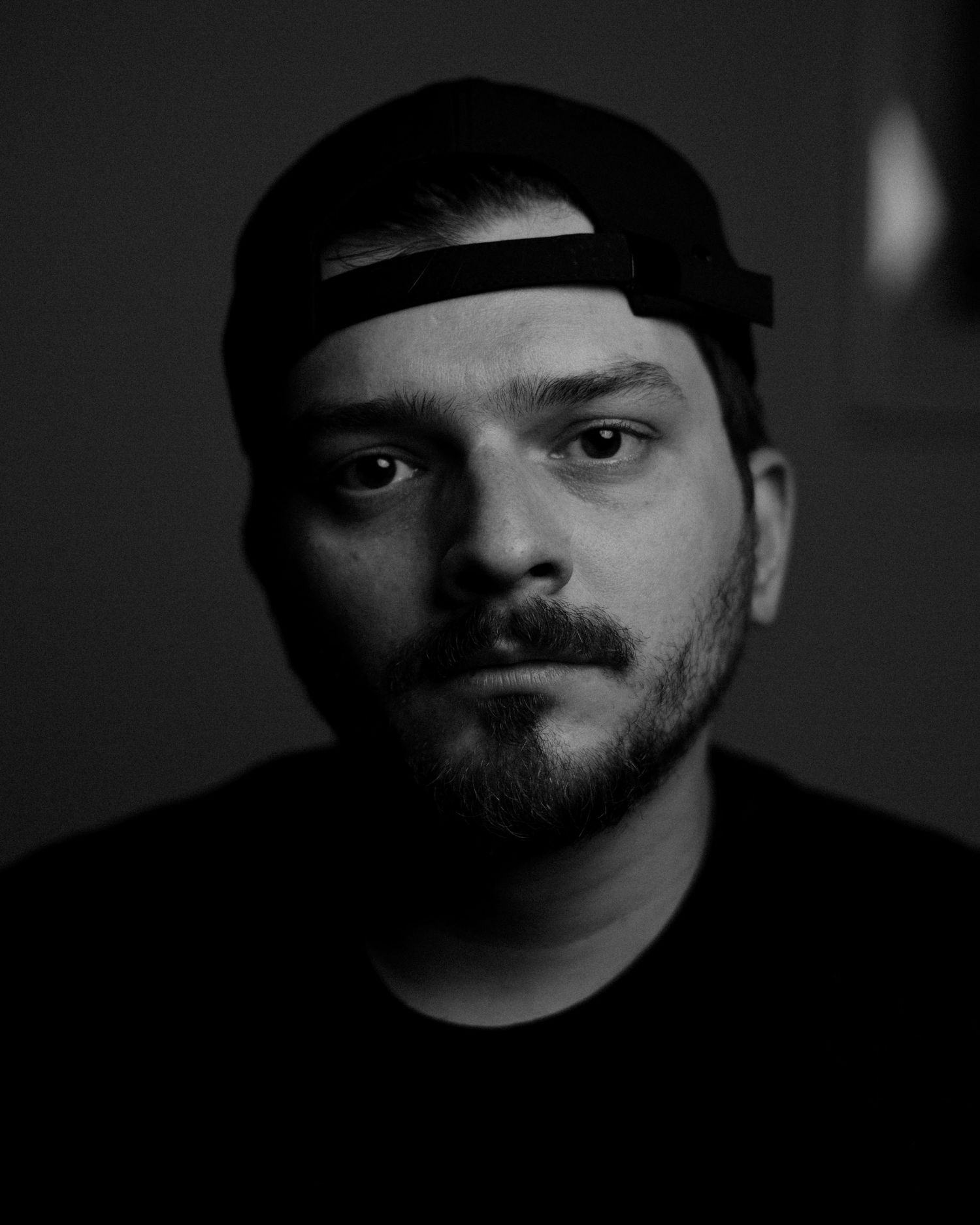 Black and white portrait in dramatic lighting showing a bearded figure wearing a backwards baseball cap.