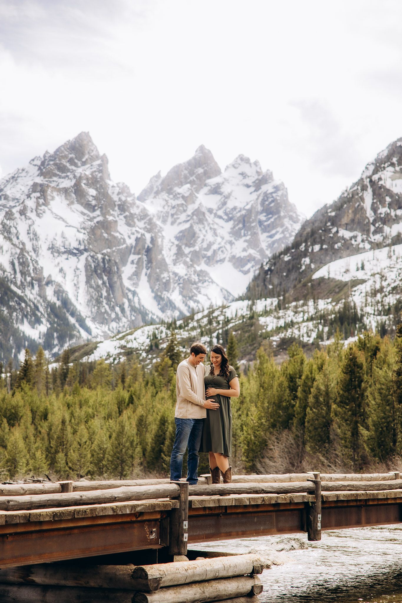 A couple stands on a wooden dock surrounded by snow-capped mountains and evergreen forest in Grand Teton National Park.