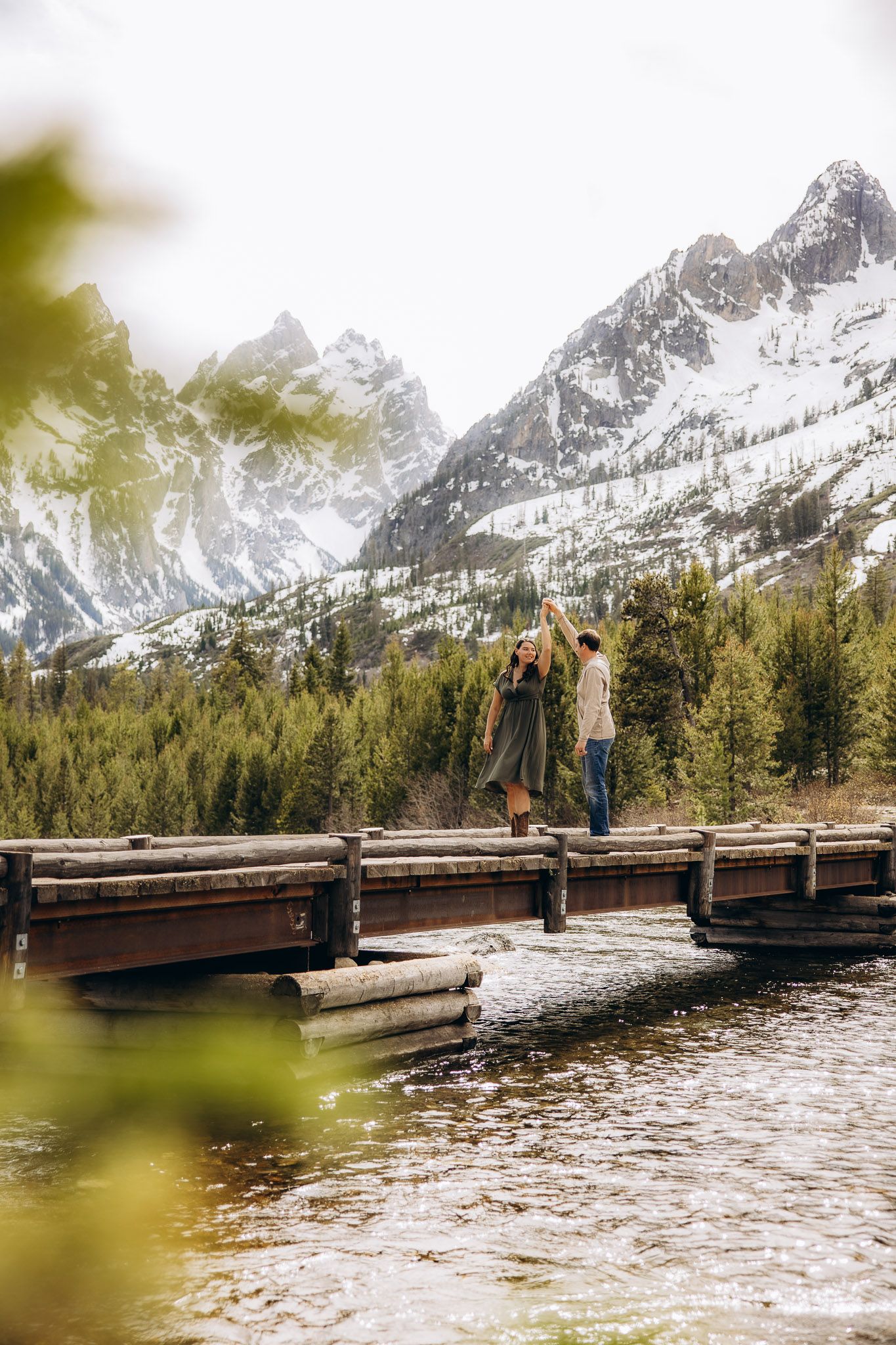 Two people enjoying a scenic wooden bridge overlook with snow-capped mountain peaks and evergreen forest in the background.