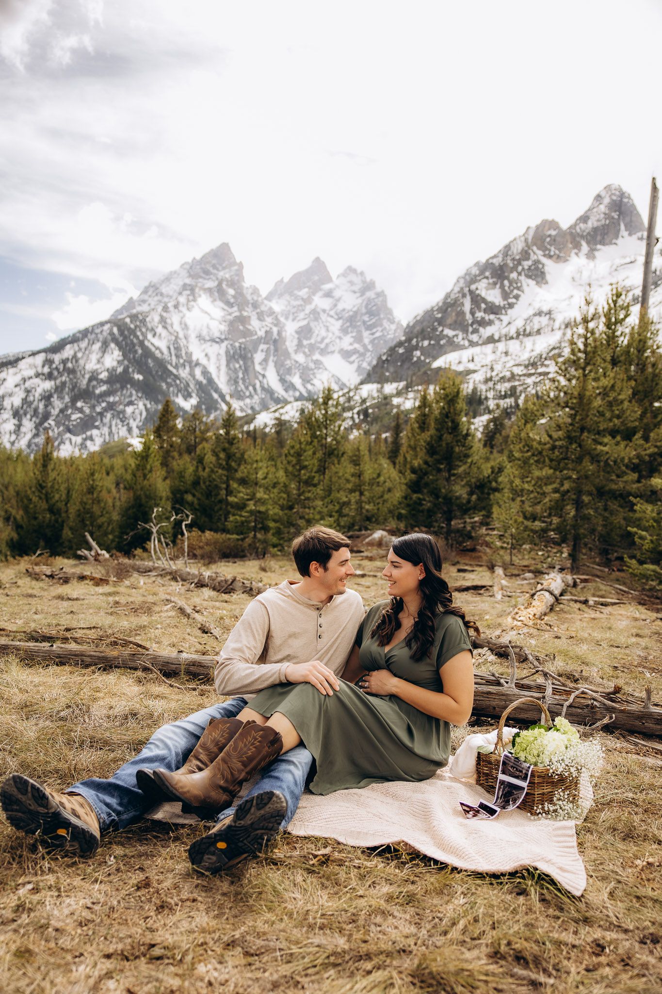 A couple enjoys a romantic picnic with snow-capped Grand Teton mountains and evergreen forest in the background.