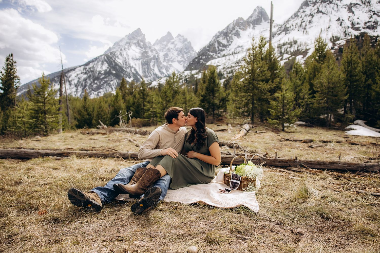 A couple enjoys a picnic on a blanket with stunning snow-capped mountains and evergreen forest in the background.