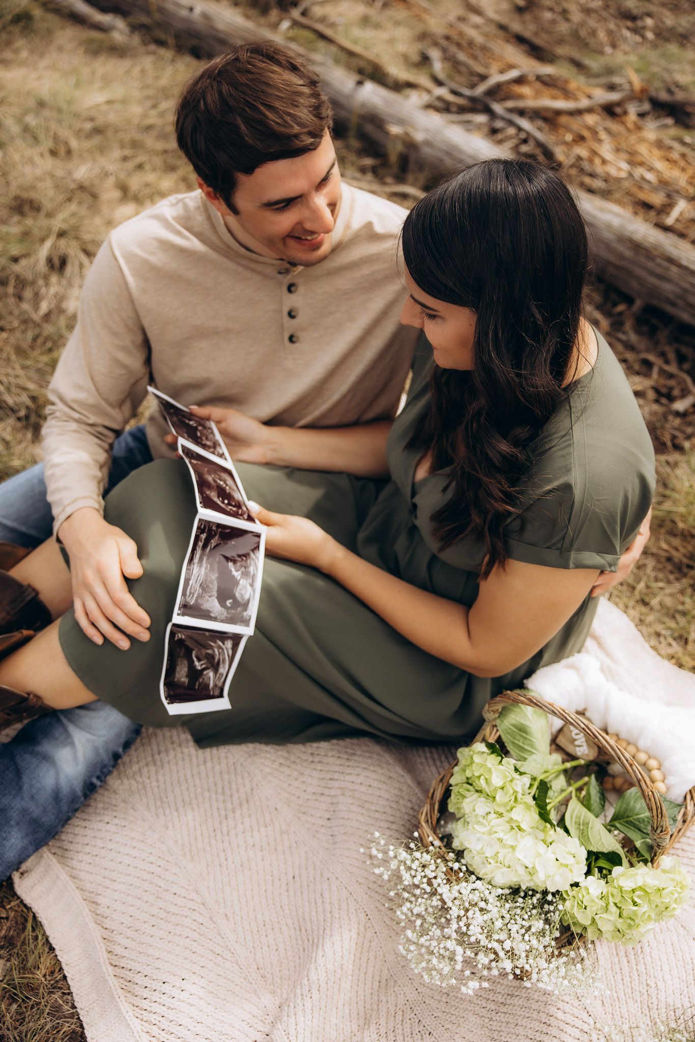 A couple sits on a blanket outdoors looking at ultrasound photos with flowers beside them.