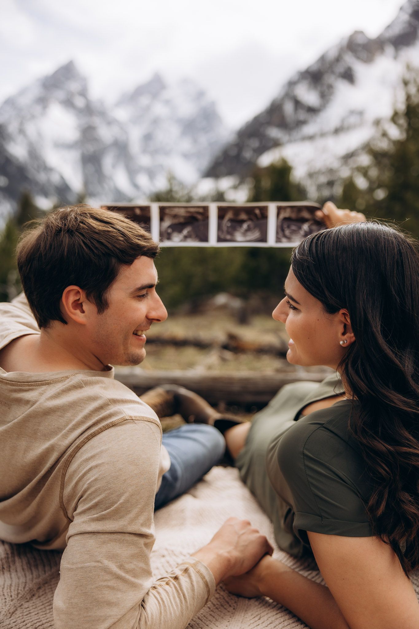 A couple shares a loving moment in nature while holding ultrasound photos against a backdrop of snow-capped mountains.
