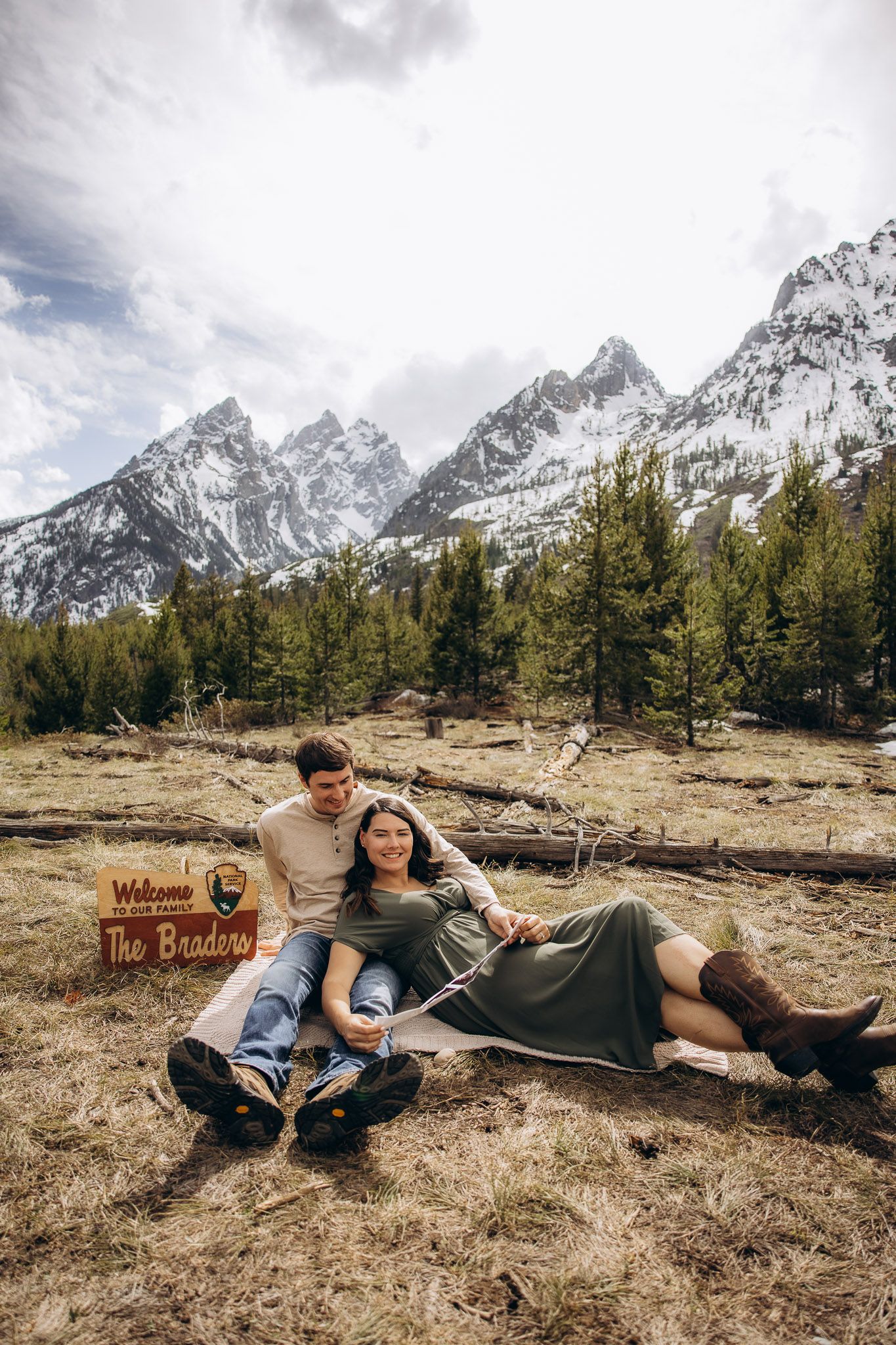 A couple relaxes on the ground near a wooden sign with snow-capped Grand Teton mountains and evergreen forest in the background.