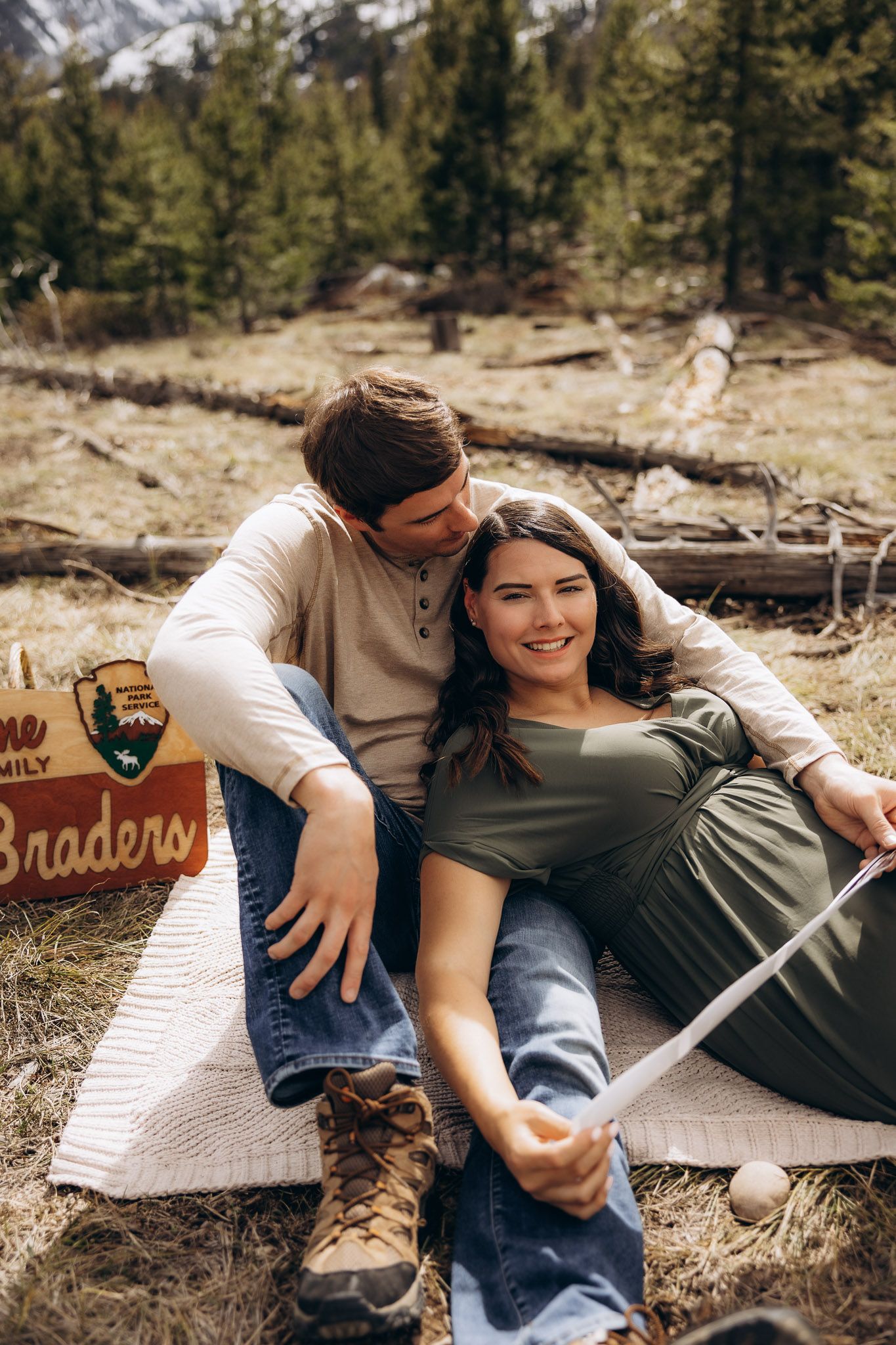 A couple relaxes together on a blanket outdoors with a wooden sign and mountainous wilderness setting.