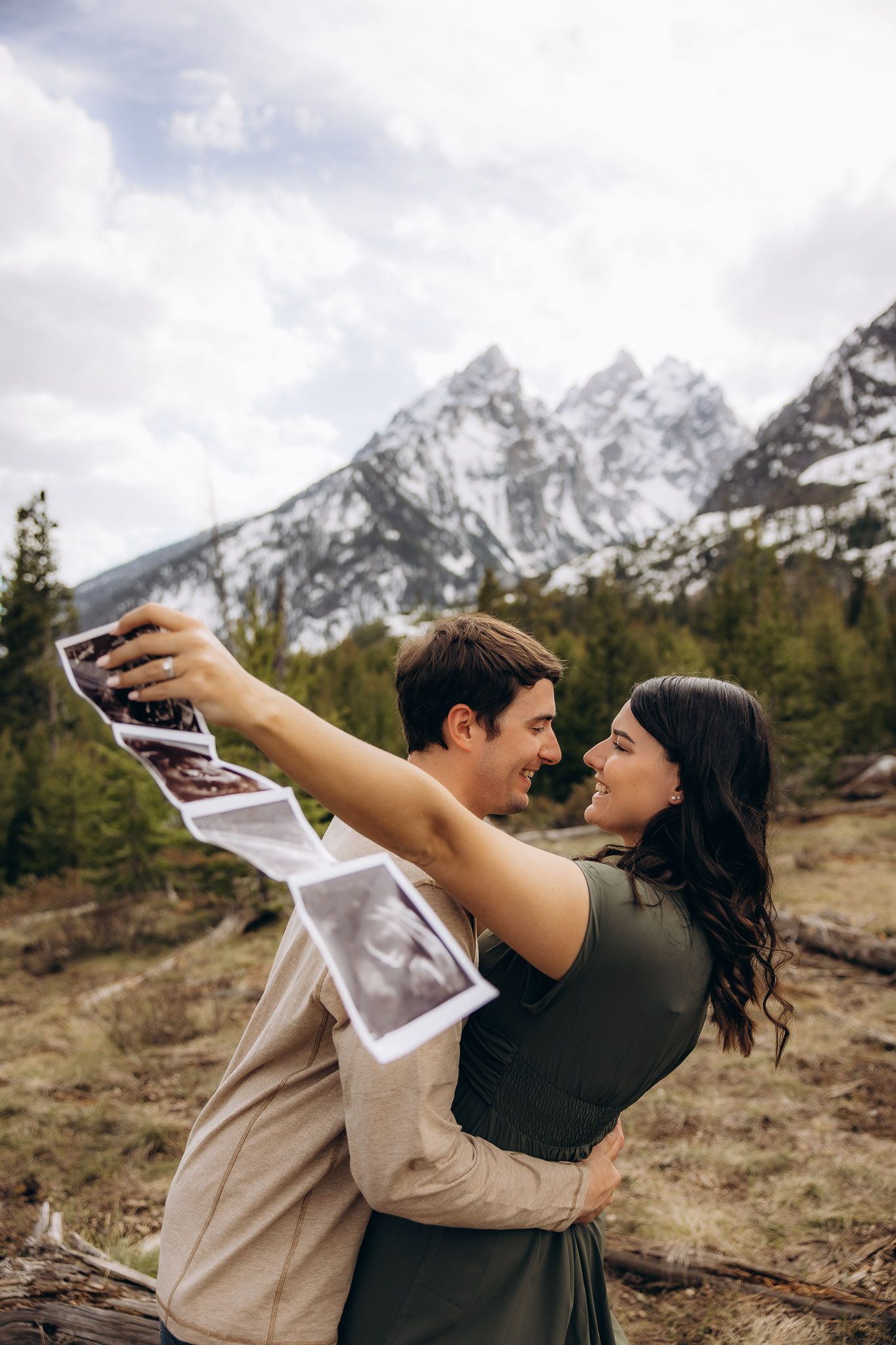 A couple sharing a romantic embrace against a backdrop of snow-capped mountain peaks and evergreen forest in autumn.