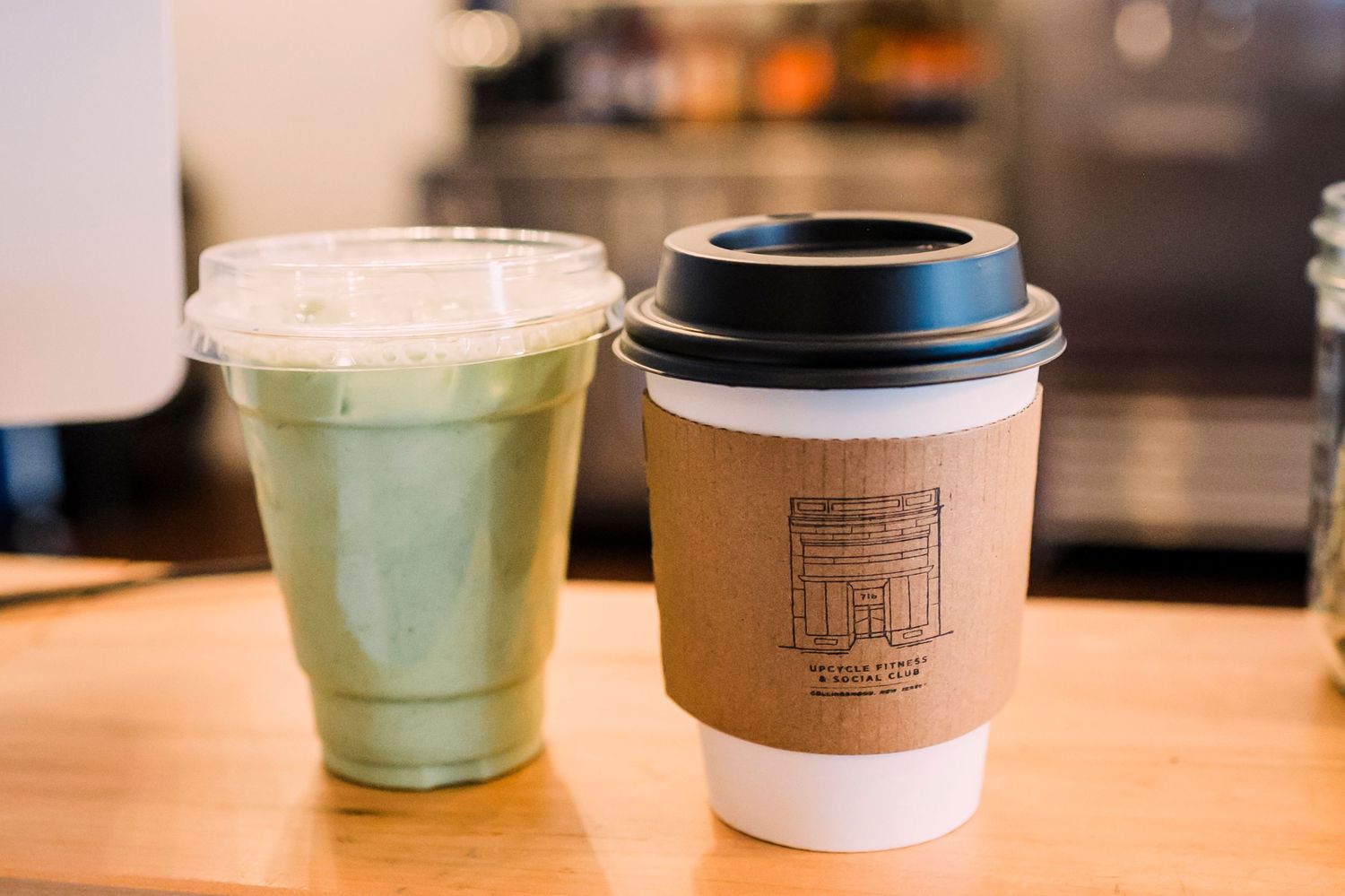 Two takeaway beverages on wooden counter - one green matcha drink in clear cup and one hot coffee in paper cup with logo.