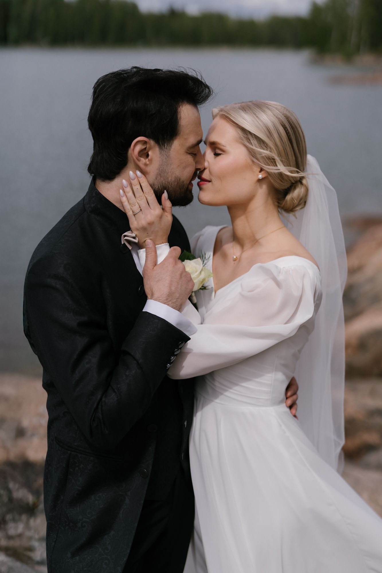Romantic wedding couple embraces by a lake, with bride in white flowing dress and groom in black suit at dusk.