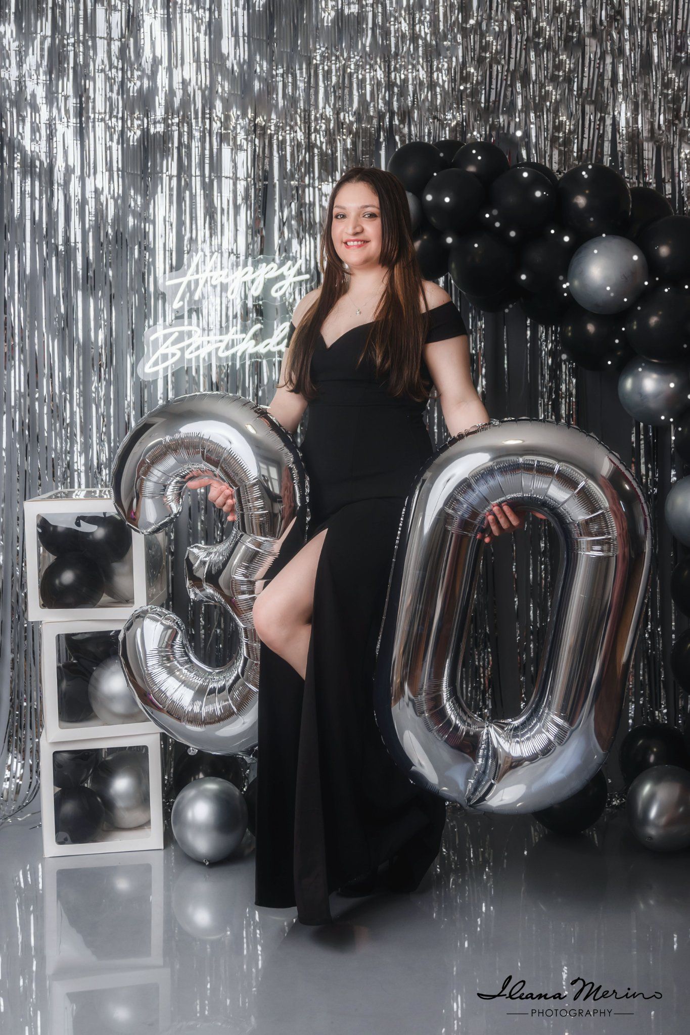 Woman in black gown posing with silver 30 balloon numbers at a glamorous birthday celebration with black and silver decor.