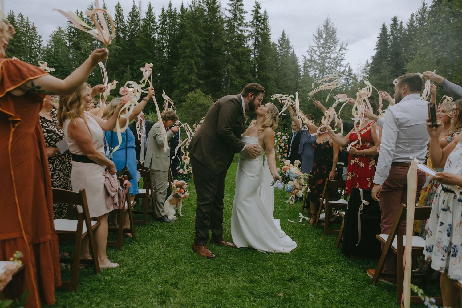 Wedding couple shares a kiss on a grassy lawn surrounded by cheering guests and evergreen trees.