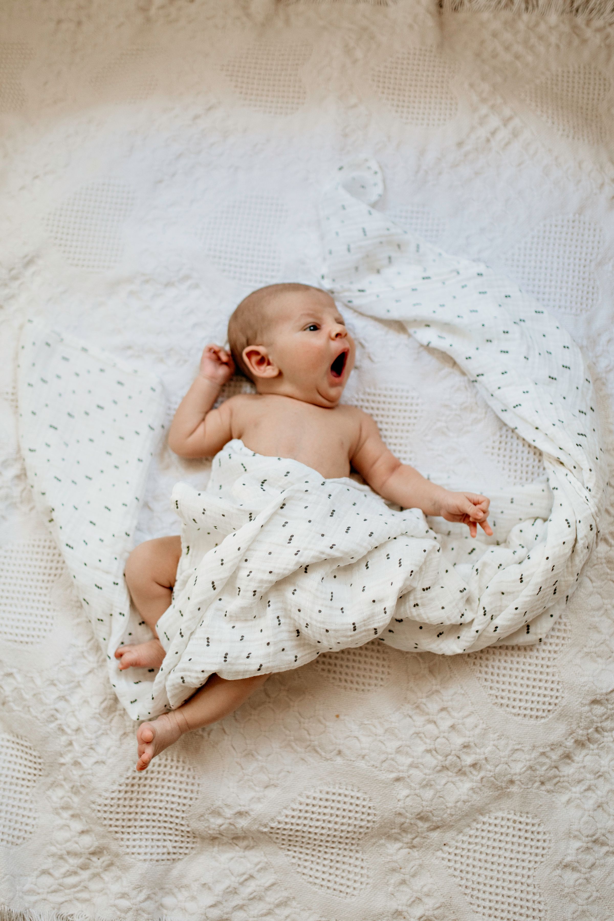 Baby lying on white textured blanket yawning while wrapped in a patterned swaddle cloth for in home photos.