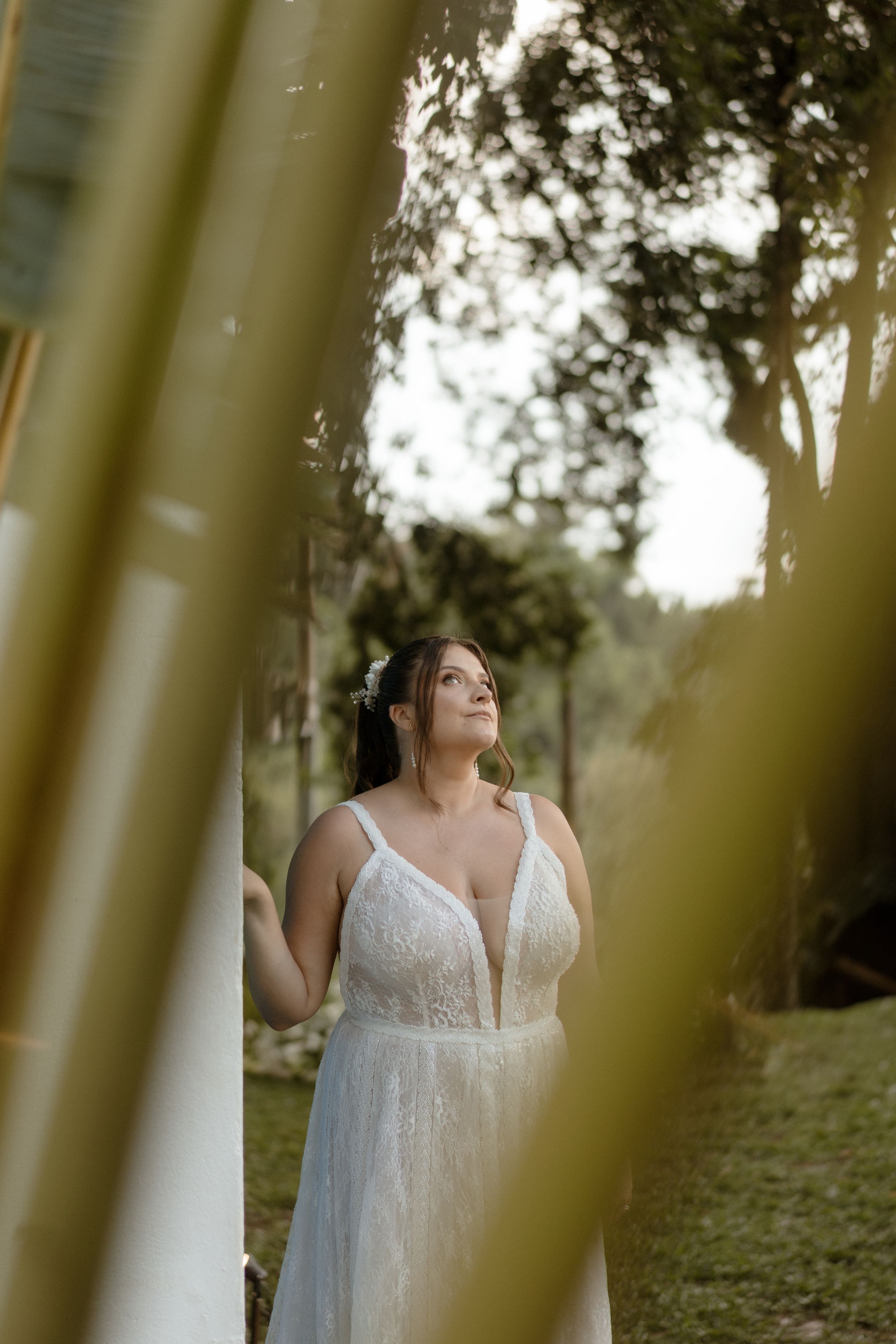 A person in a white lace wedding dress poses in a natural outdoor setting with soft lighting filtering through trees.