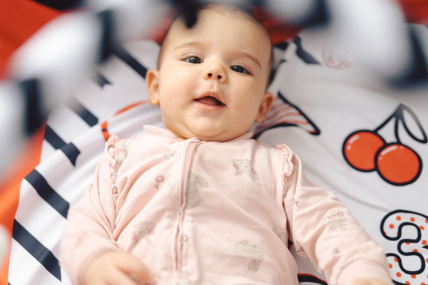 Baby wearing light pink top lies on striped blanket with cherry print pattern.