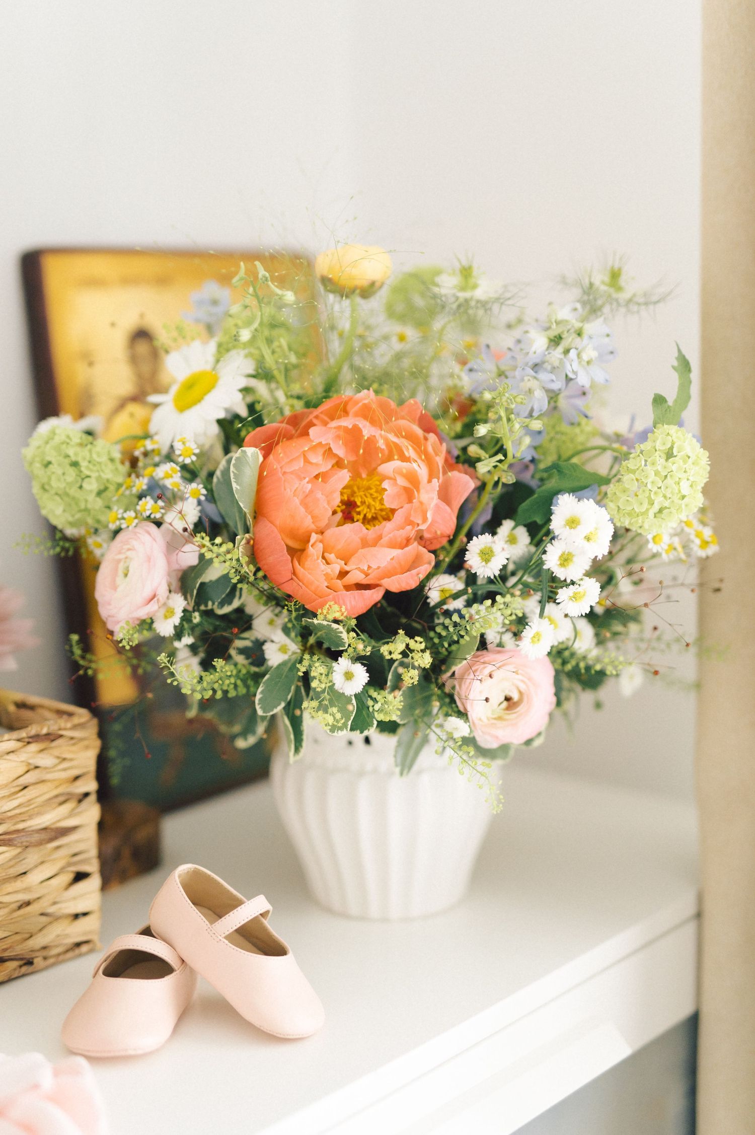 Delicate floral arrangement with coral peonies and daisies in white vase next to pale pink ballet shoes on white shelf.