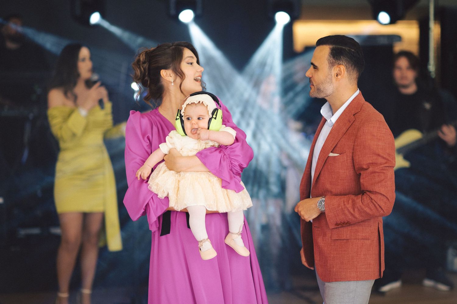 People interact on a stage with bright spotlights while someone holds a baby wearing headphones during a televised show.