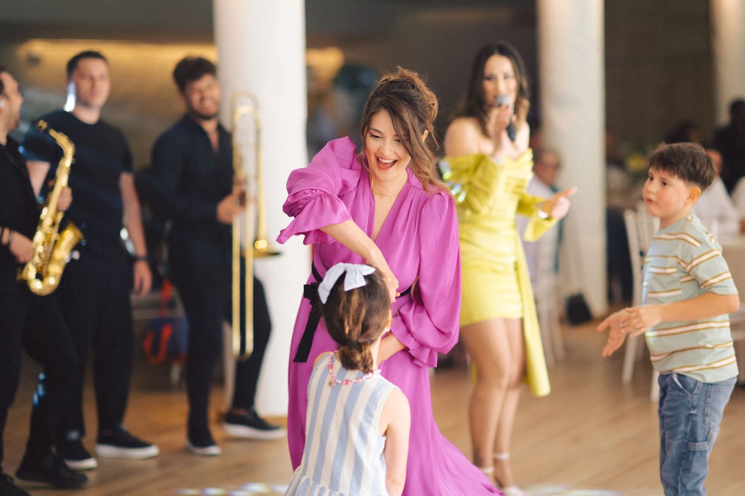 People dance and celebrate at a party with musicians playing saxophone in the background while dressed in colorful attire.
