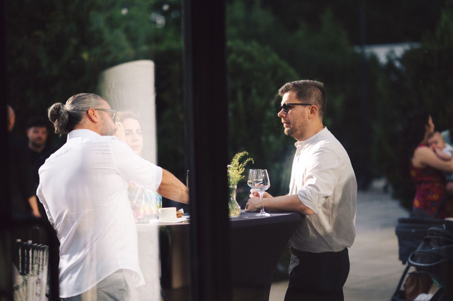 Two waiters in white shirts having a conversation while holding wine glasses at an outdoor restaurant venue.