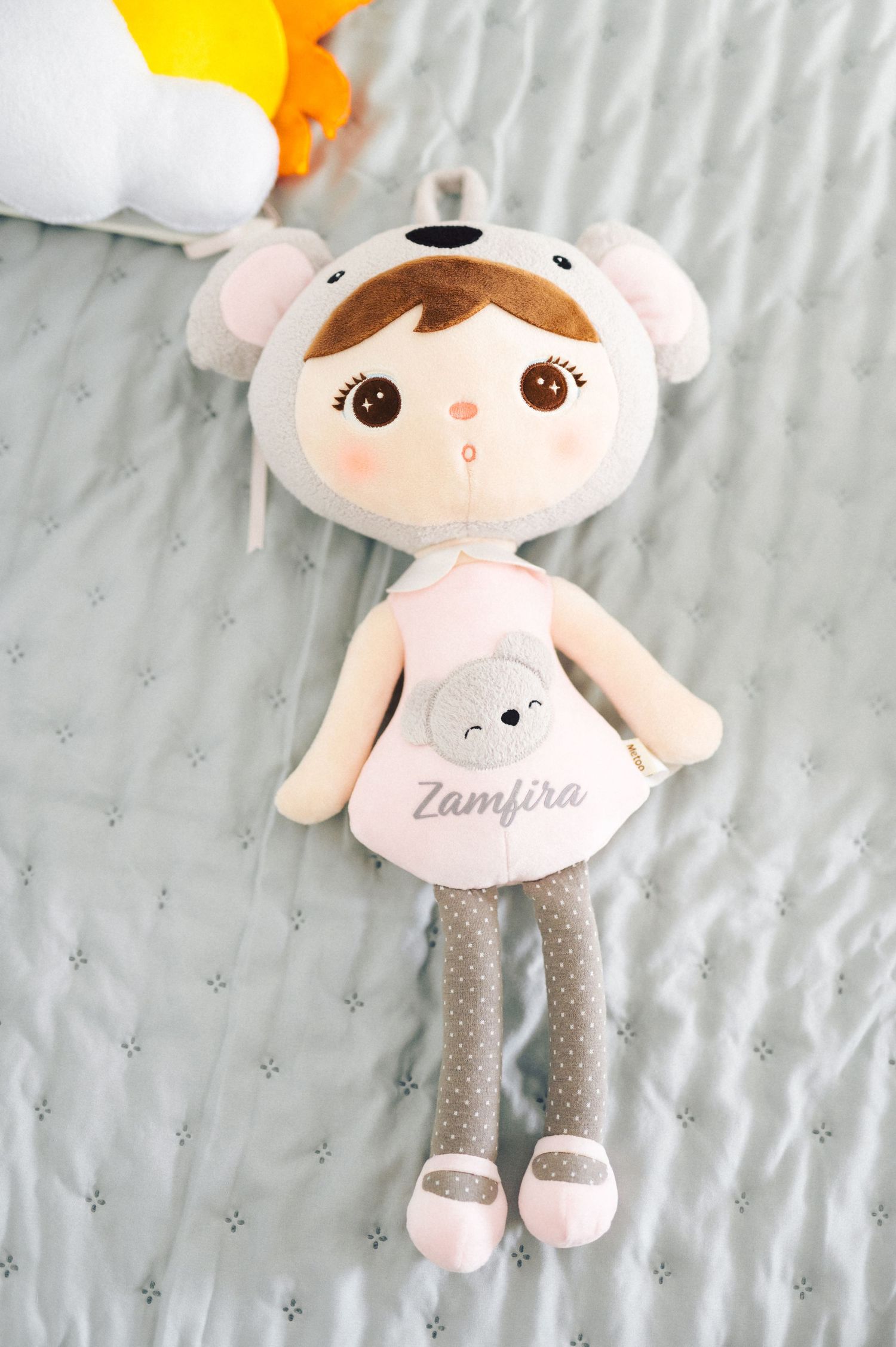 Soft plush doll in pink dress wearing white animal ears and grey polka dot leggings on grey bedding.