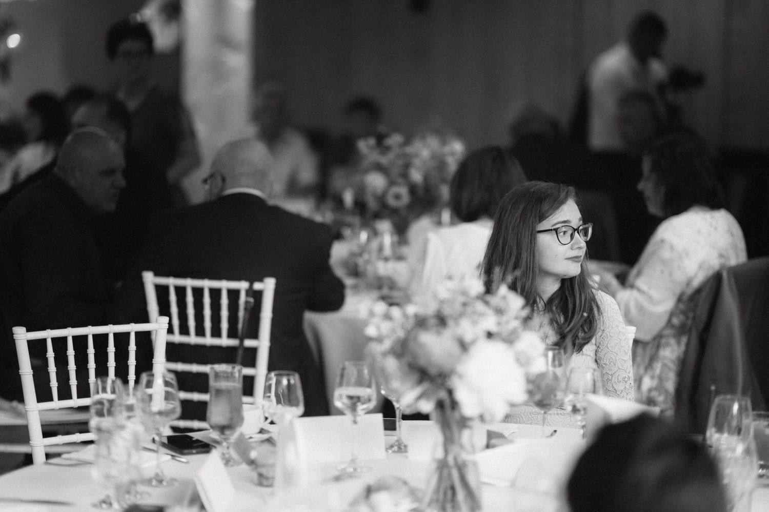 A black and white photo of wedding reception guests seated at decorated tables with white chiavari chairs.