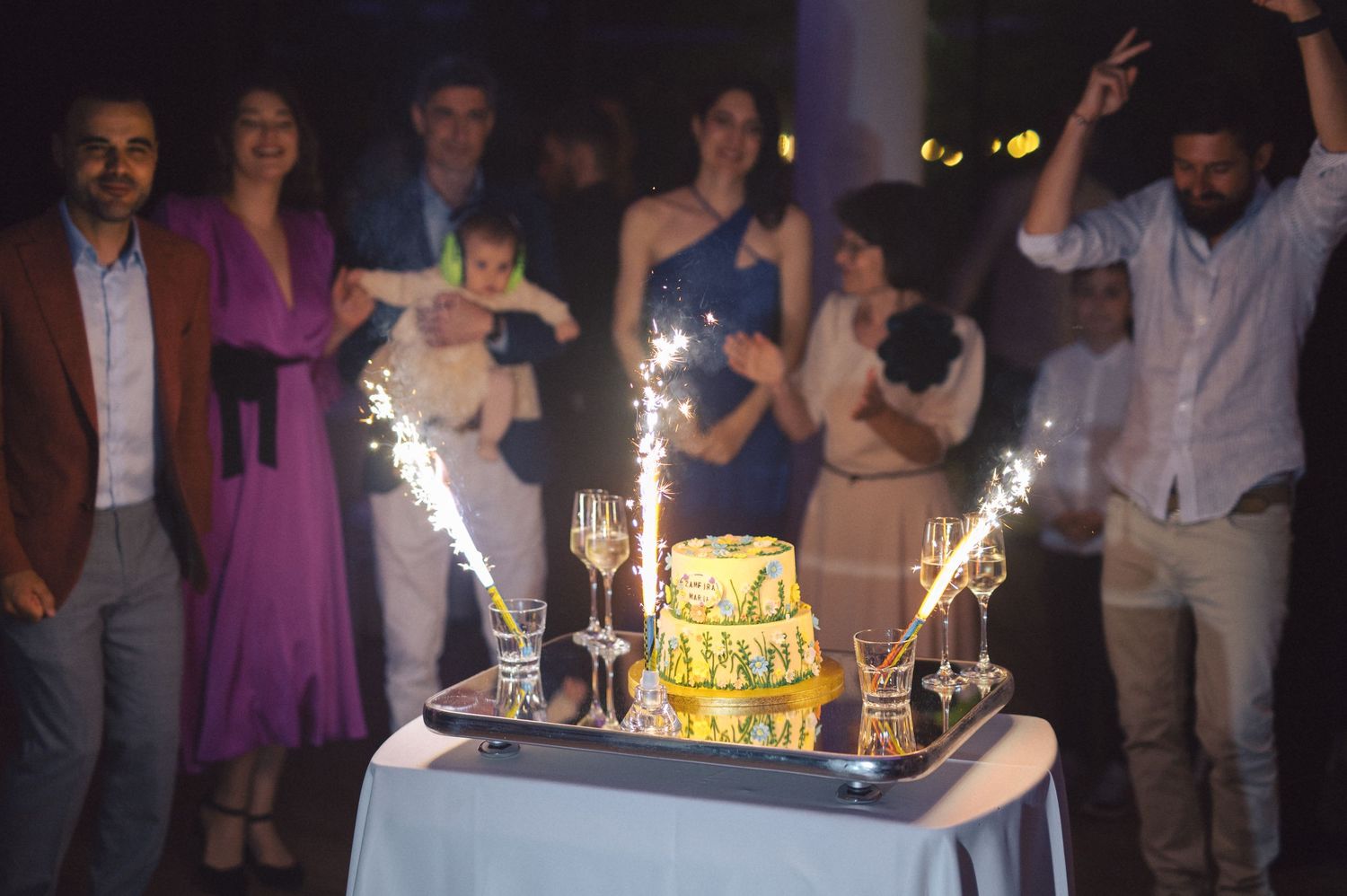 A yellow celebration cake with sparklers sits on a table at a nighttime party as guests celebrate in the background.
