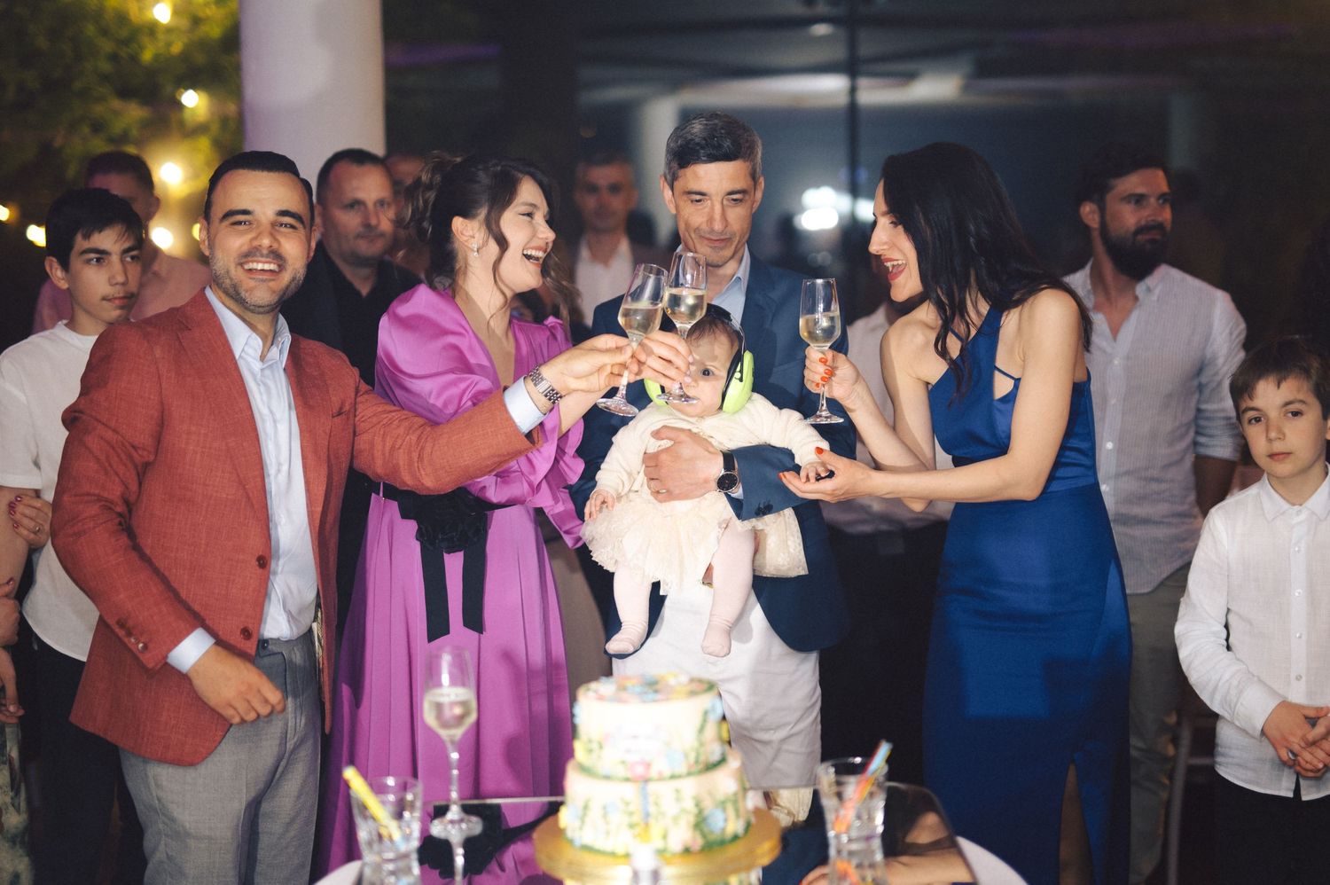 A group gathers around a white wedding cake at an evening celebration while making a toast.