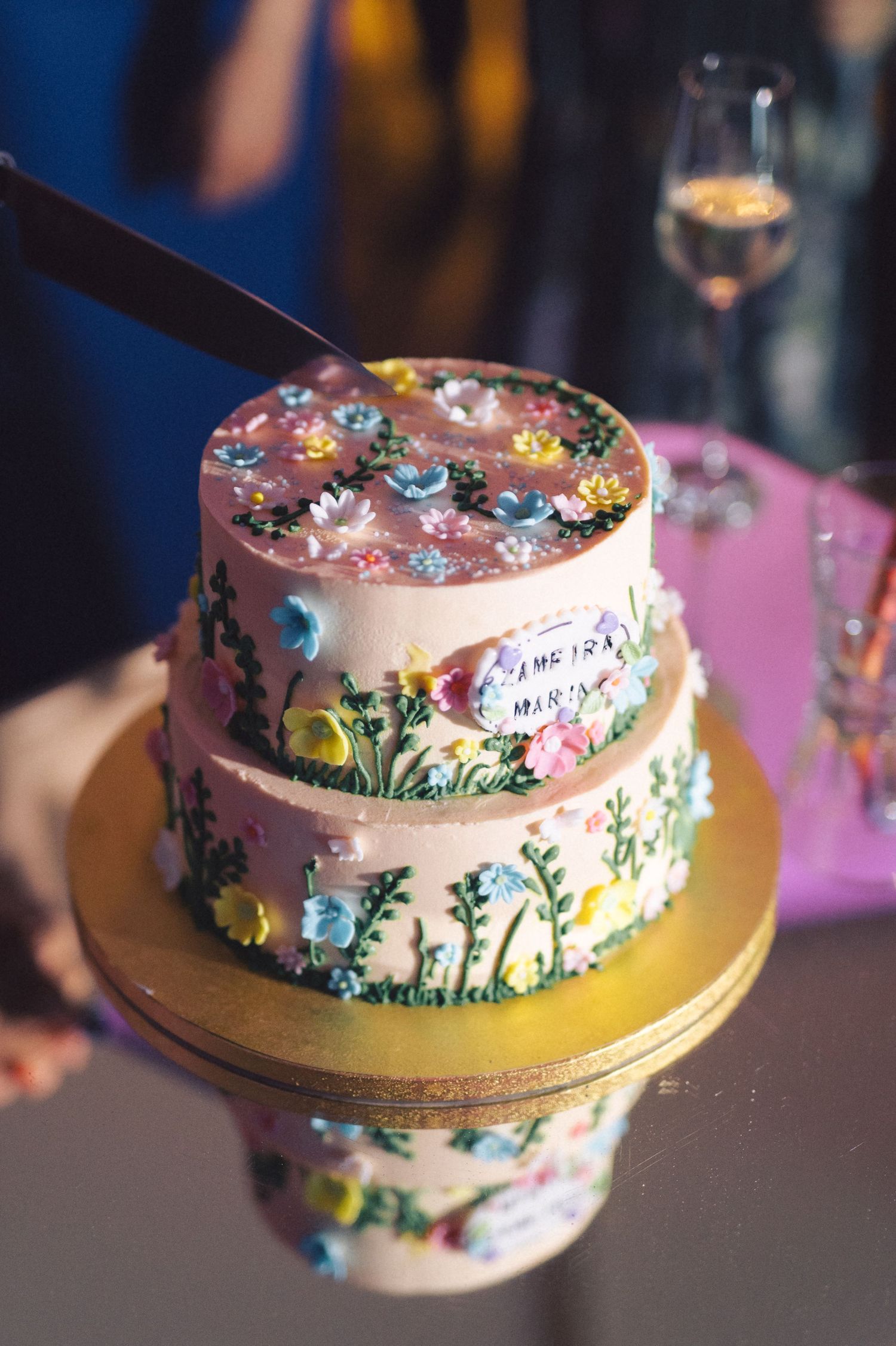 A two-tiered pink birthday cake decorated with colorful floral designs and butterflies on a gold cake stand.
