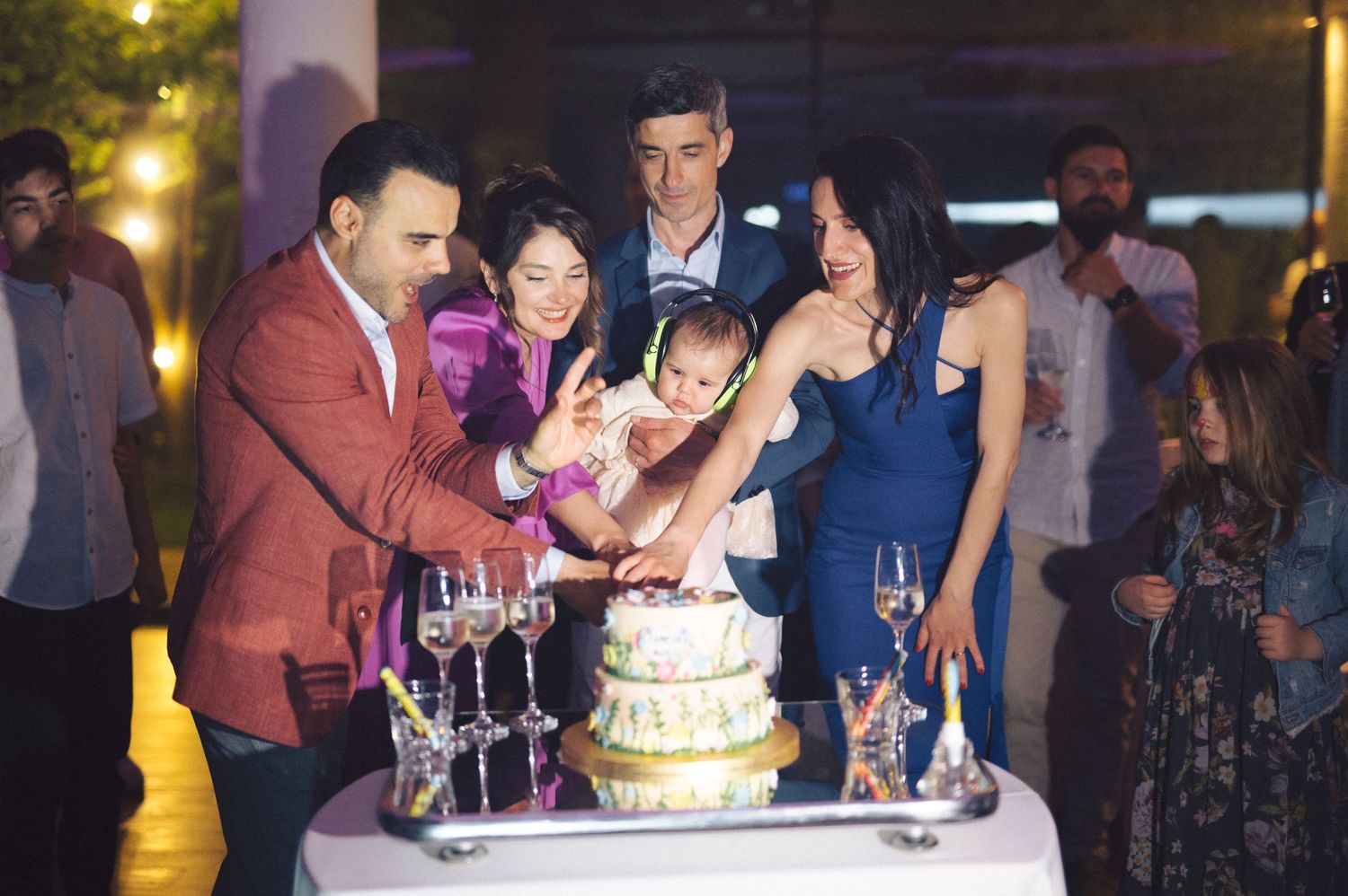 A group of evening party guests gather around a decorated cake with champagne flutes at an elegant celebration.