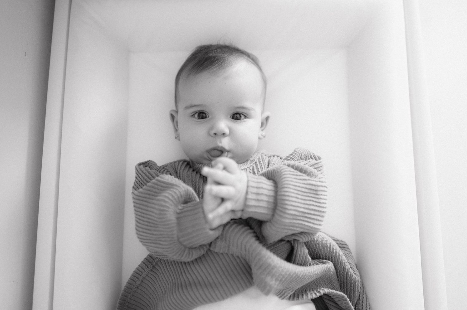 Black and white photo of a baby wearing a striped outfit while seated indoors against a white background.