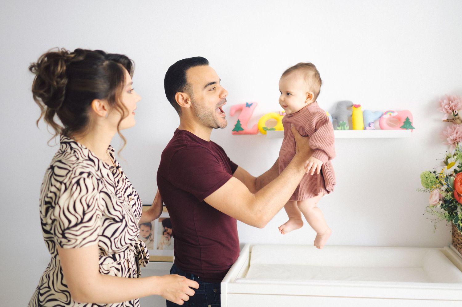 Parents playing and interacting with their baby in a bright nursery room with floating shelves and decorative items.
