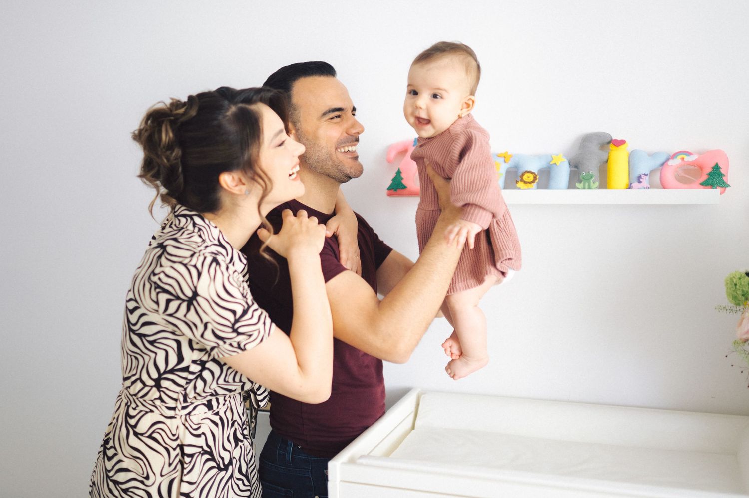 A couple interacts playfully with their young baby while standing in a bright room with shelving in the background.
