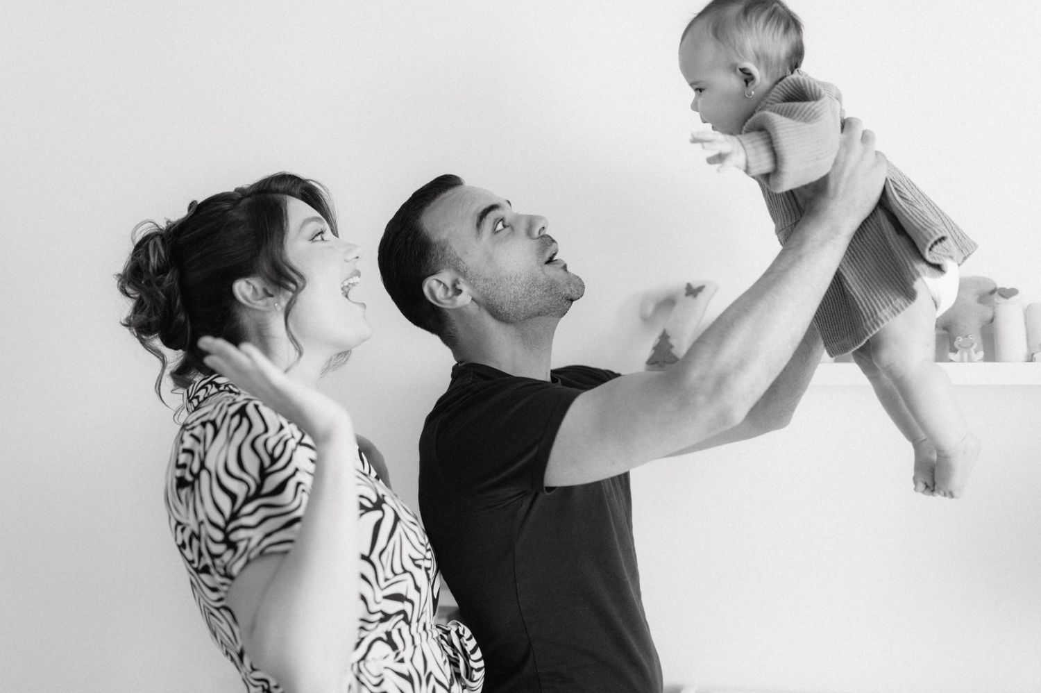 A joyful black and white family portrait shows parents smiling up at their baby being lifted in the air.