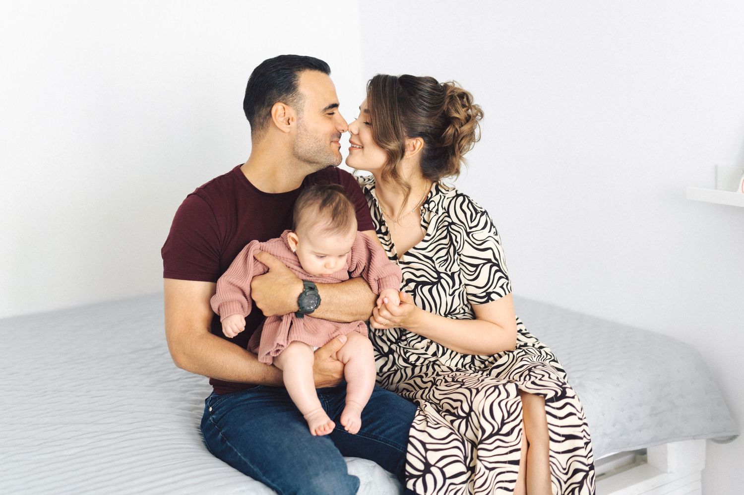 Happy parents share a tender moment with their baby on a cozy grey bed.