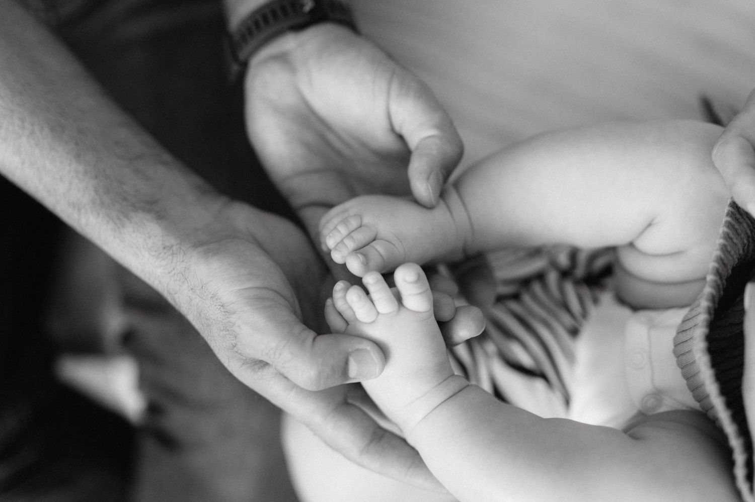 Black and white close-up of adult hands gently holding tiny baby feet against light background.