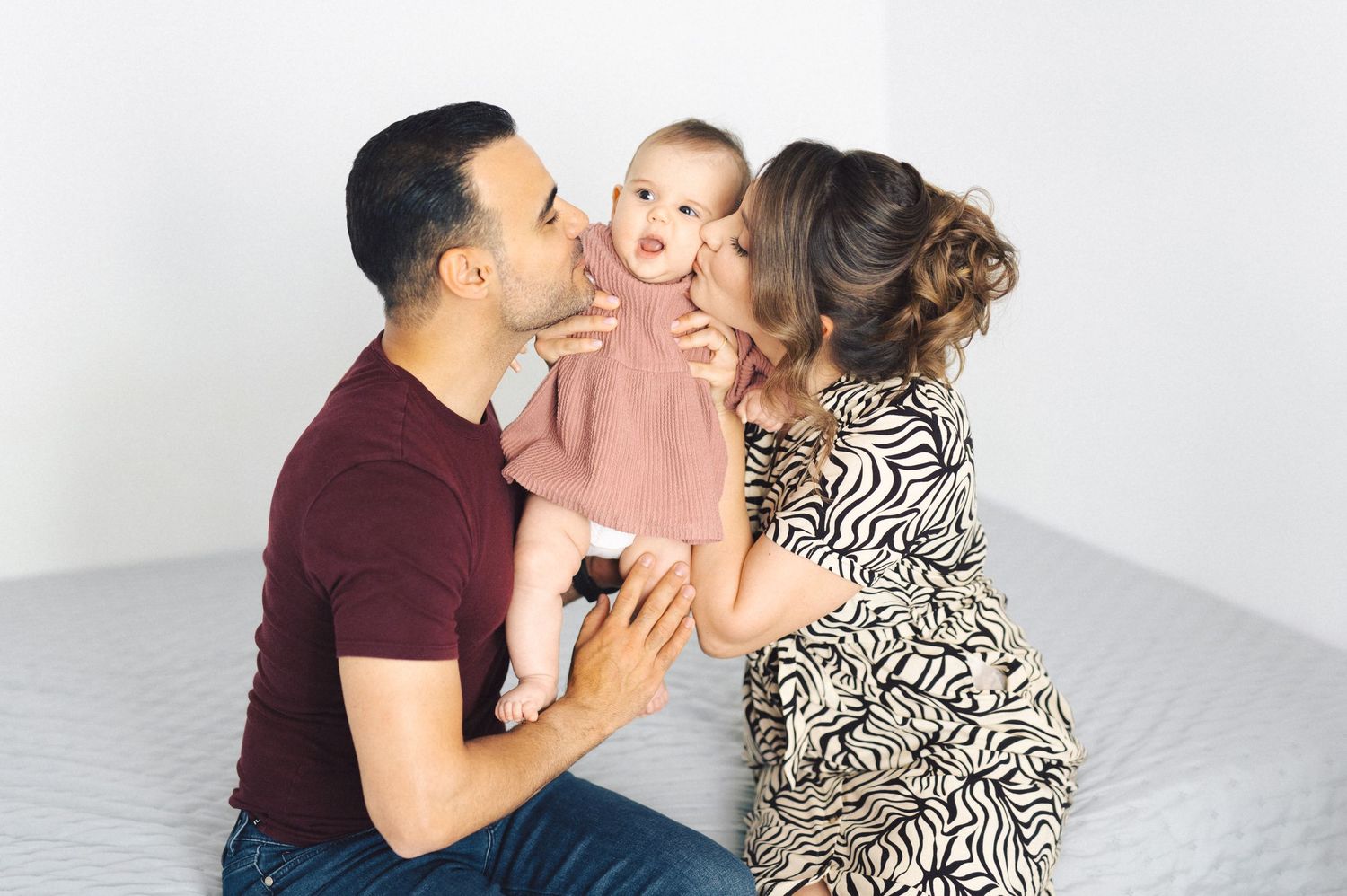 Sweet family moment with parents kissing their happy baby in pink outfit against white background.