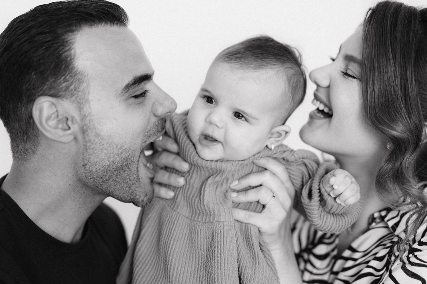 Loving family portrait in black and white showing parents with baby in knit sweater sharing joyful moment together.