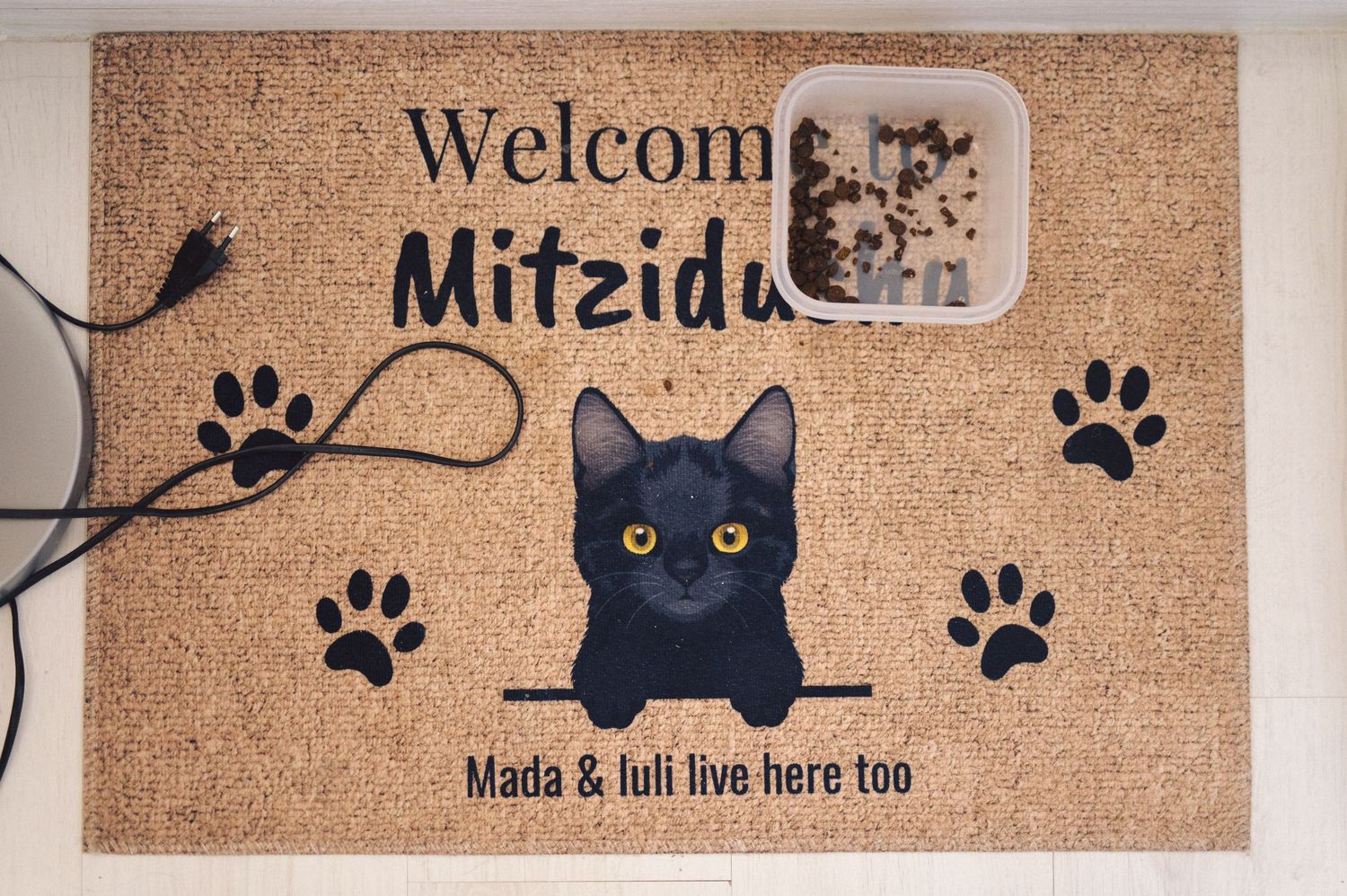 A brown doormat with a black cat design and paw prints features text reading 'Welcome Mitzida' and 'Nada & luli live here too'.