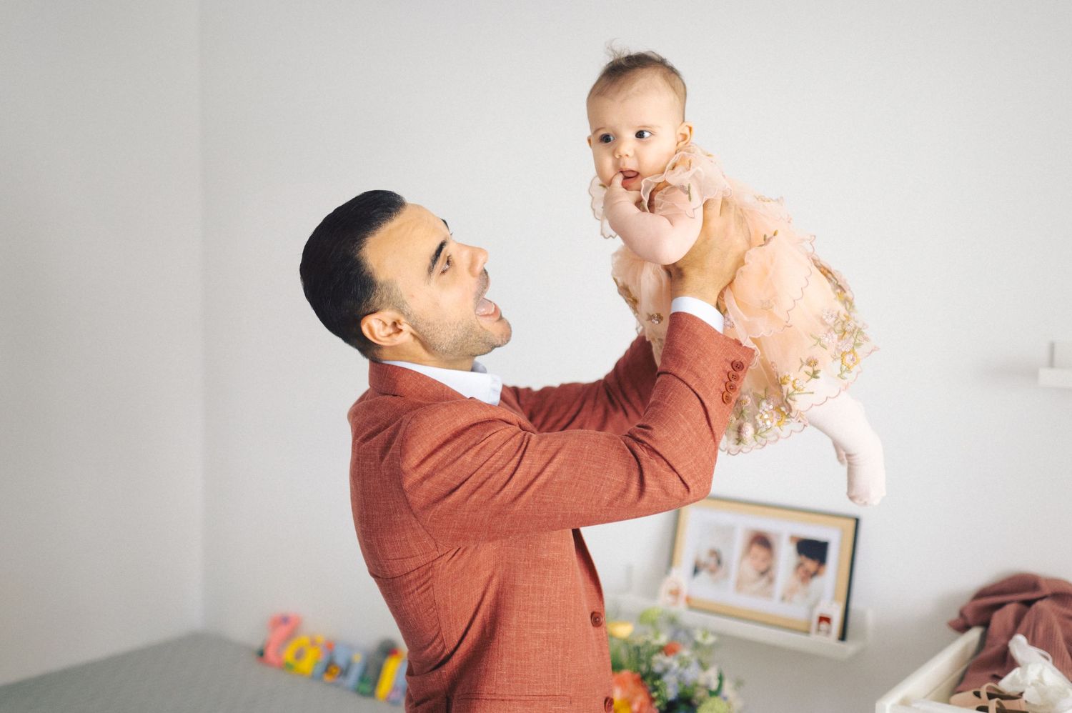A person in a rust-colored sweater lifts a joyful baby wearing pink against a light gray background.