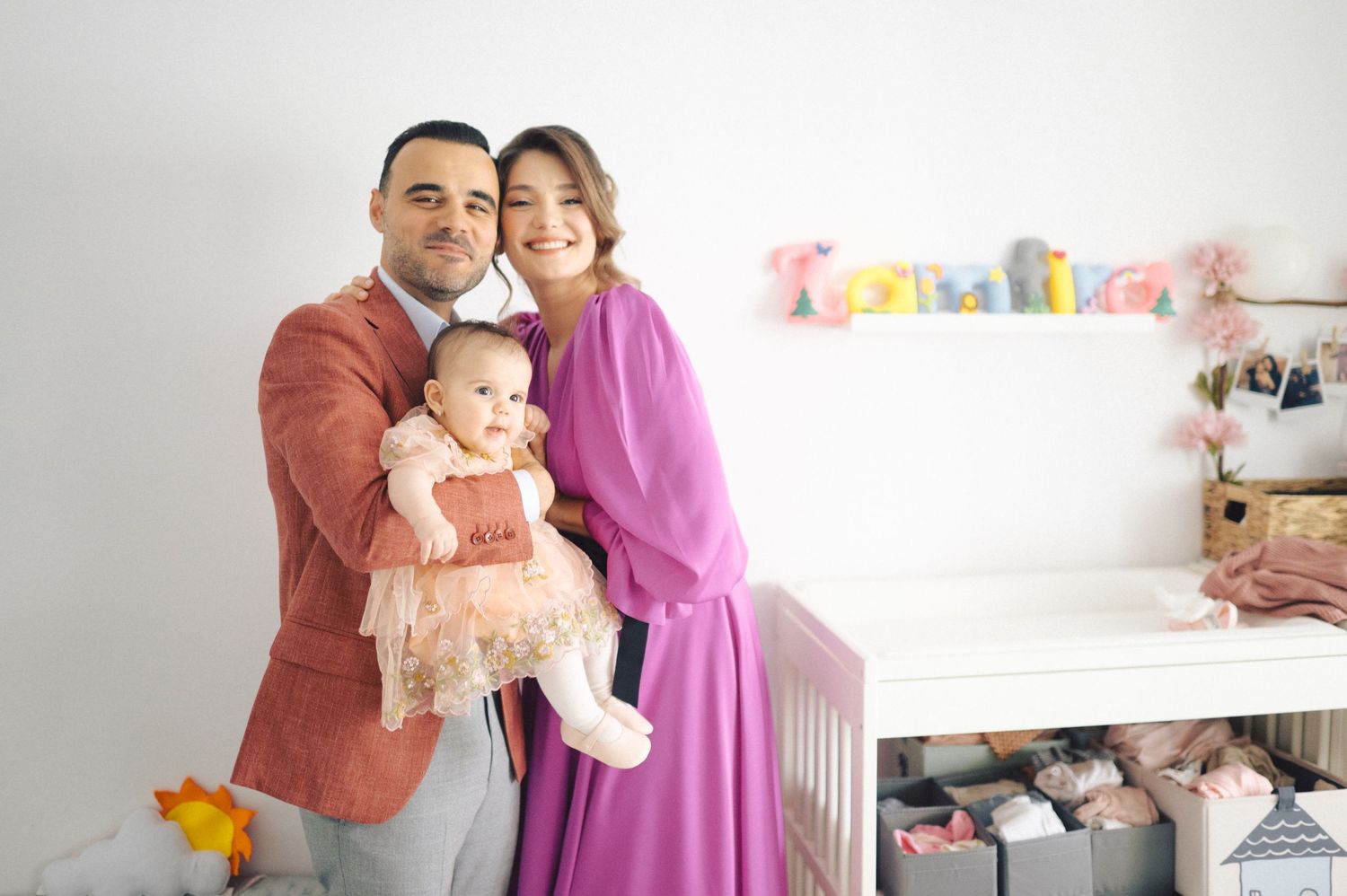A family poses together in a nursery room with white walls and colorful decorations on the shelf.