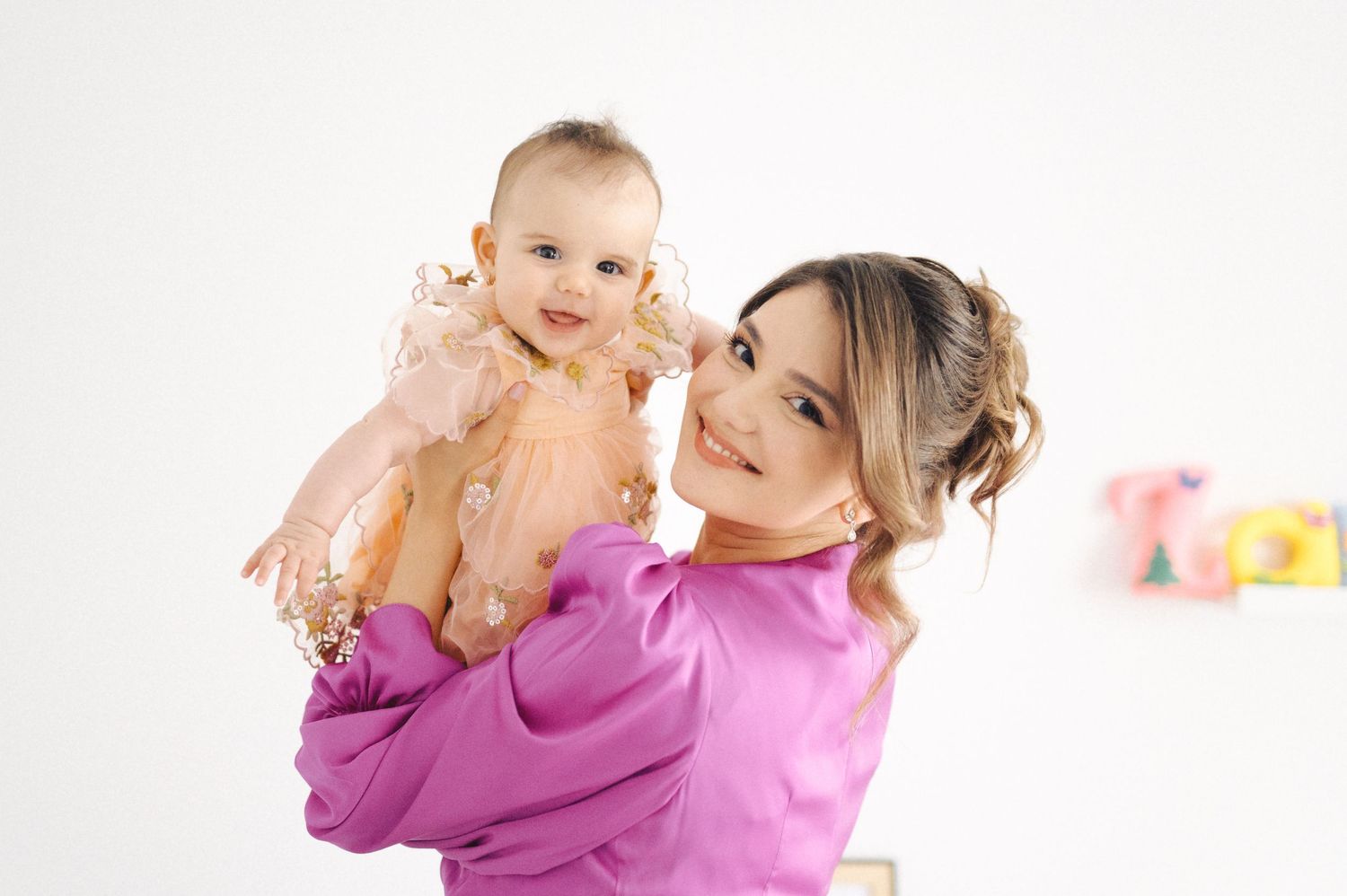 A caregiver in a bright pink sweater holds up a baby in casual attire against a white studio backdrop.