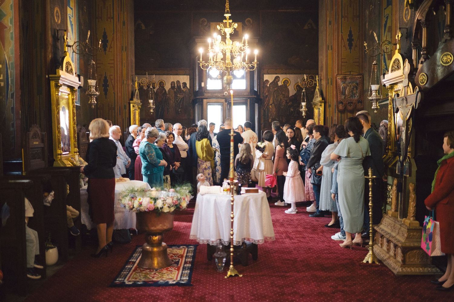 A traditional church baptism ceremony with guests gathered under a chandelier on red carpeting.
