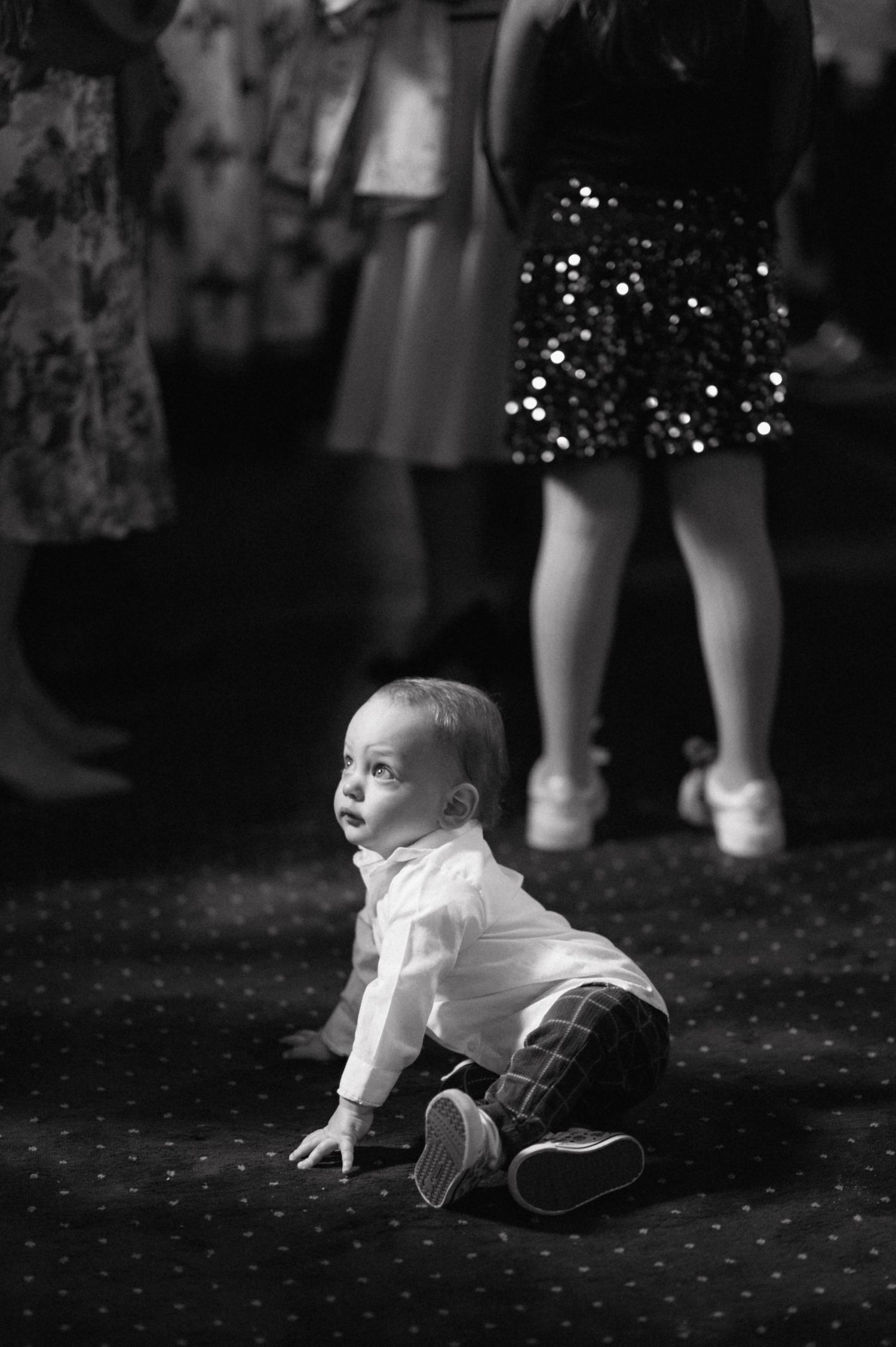 Baby in white shirt and dark pants sits on spotted carpet as someone in sparkly skirt stands in background.