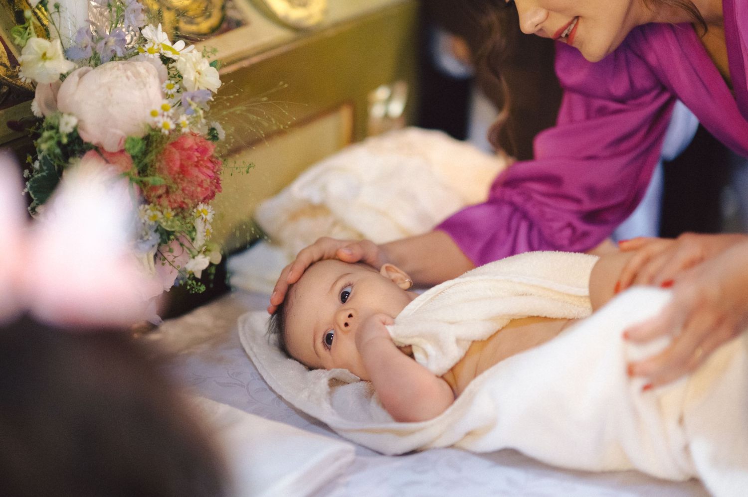 A baby in white baptismal clothing lies on a pillow surrounded by floral decorations during a christening ceremony.