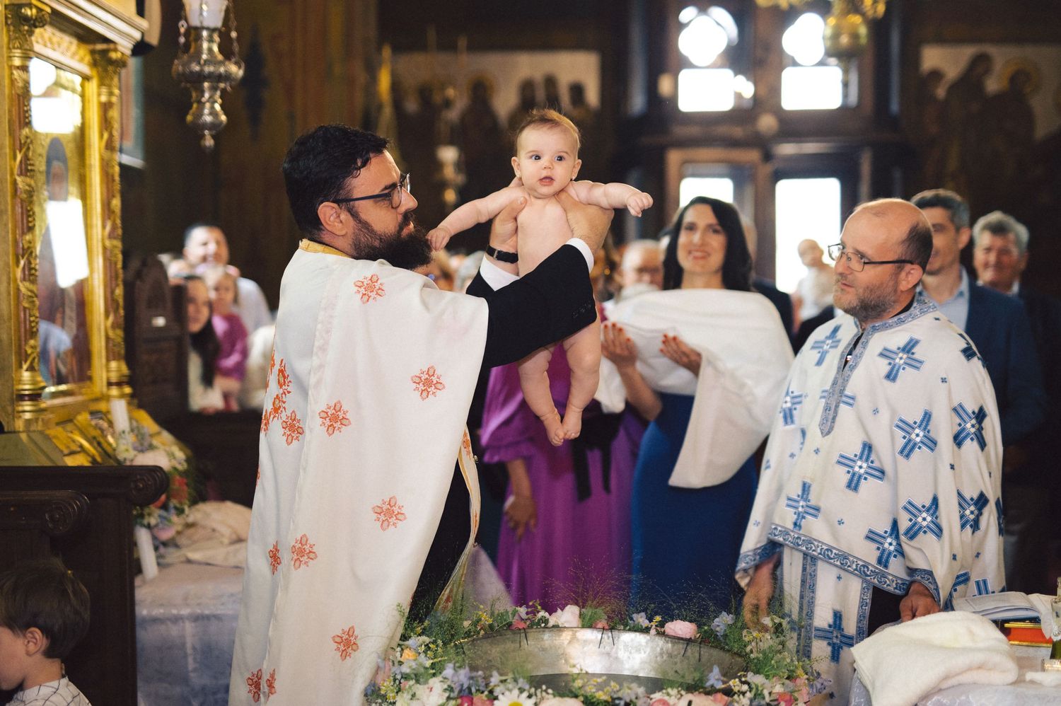 An Orthodox Christian baptism ceremony taking place inside a church with priests in traditional white robes performing the ritual.