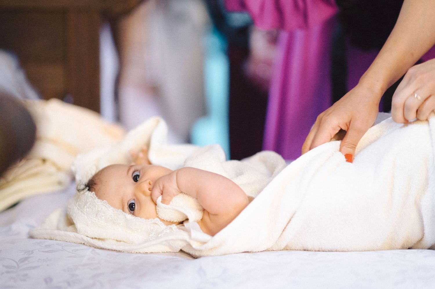 Baby lying on soft white blanket with pink garments visible in background.