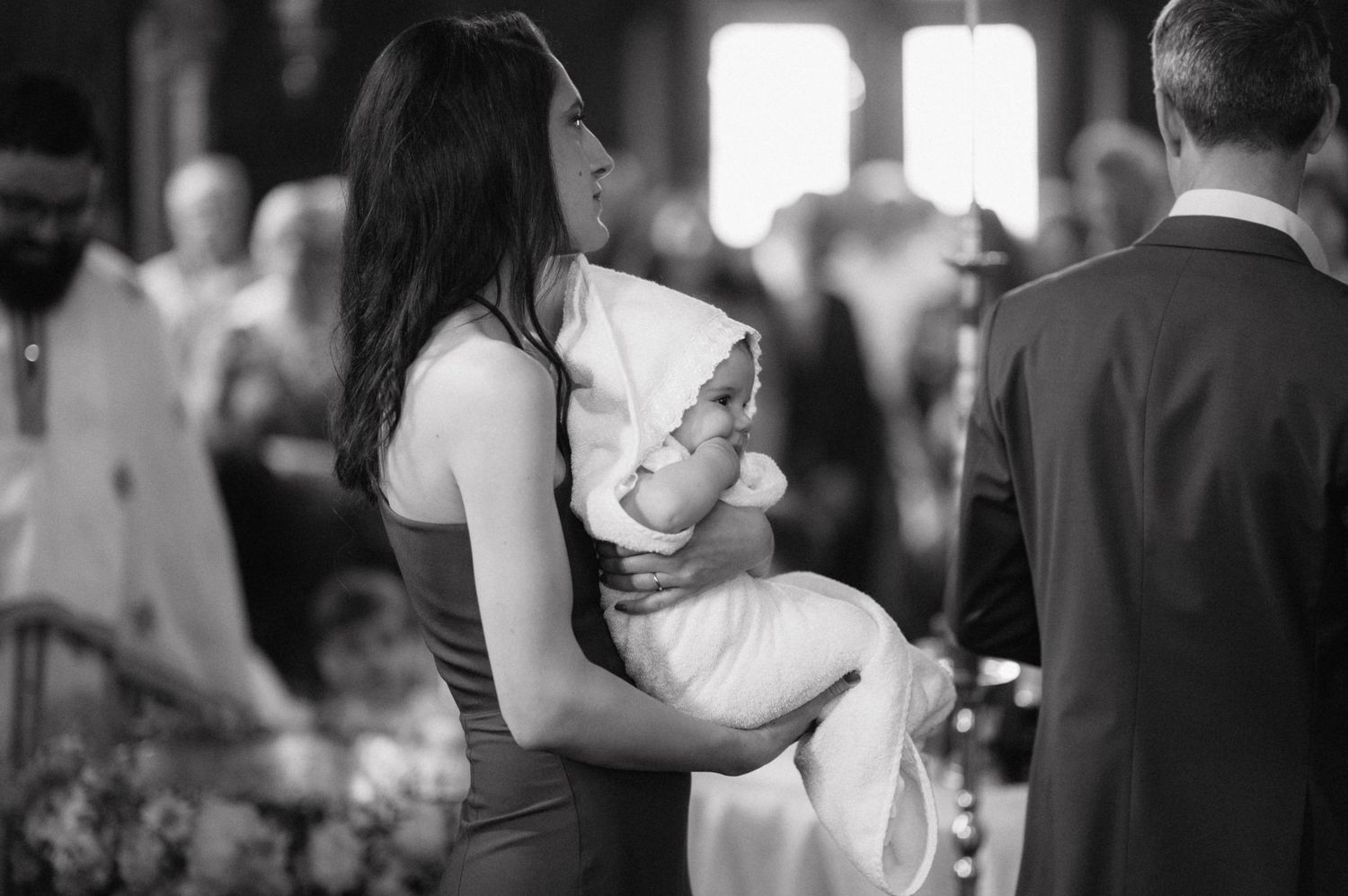A woman in a dark dress holds a baby wrapped in white during what appears to be a christening ceremony.