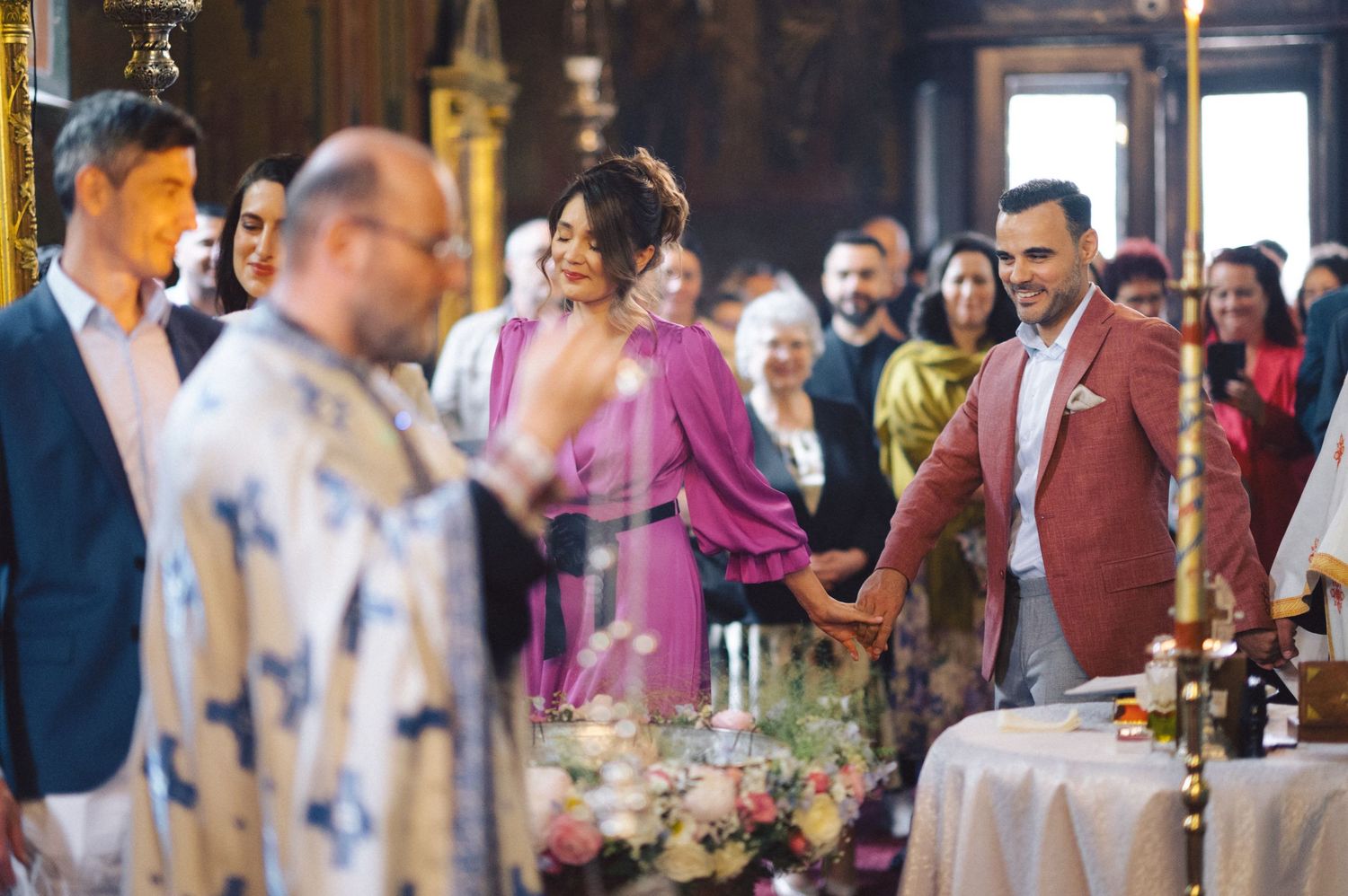 Wedding guests gather around a decorated table during an Orthodox Christian church ceremony with traditional liturgical elements.