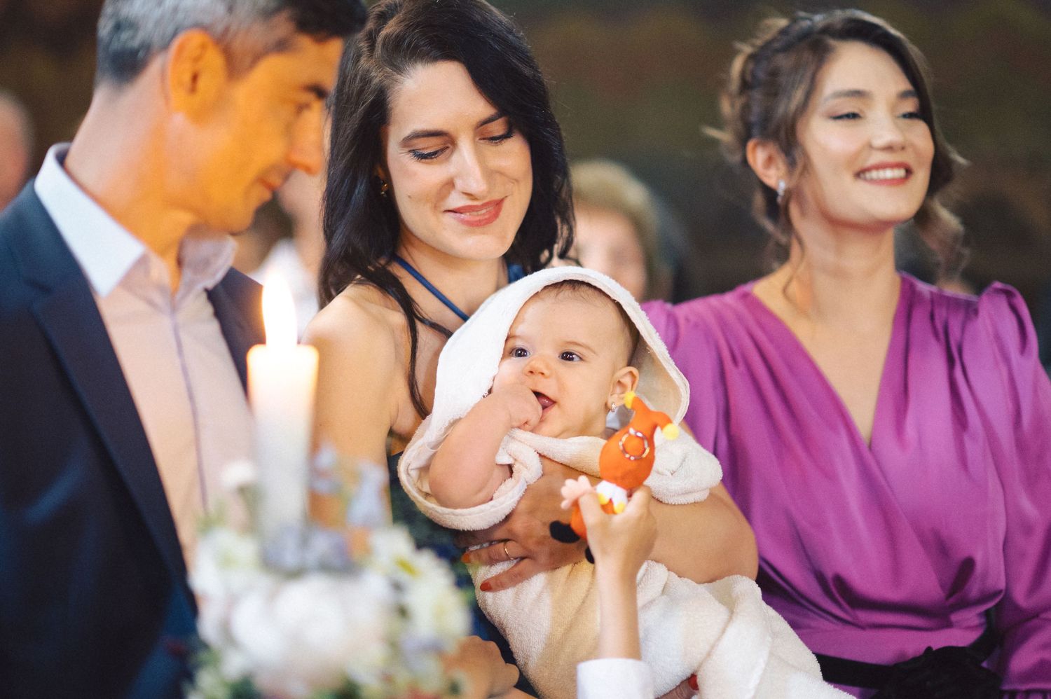 A baby in white clothing holds an orange toy while surrounded by smiling adults at a gathering.